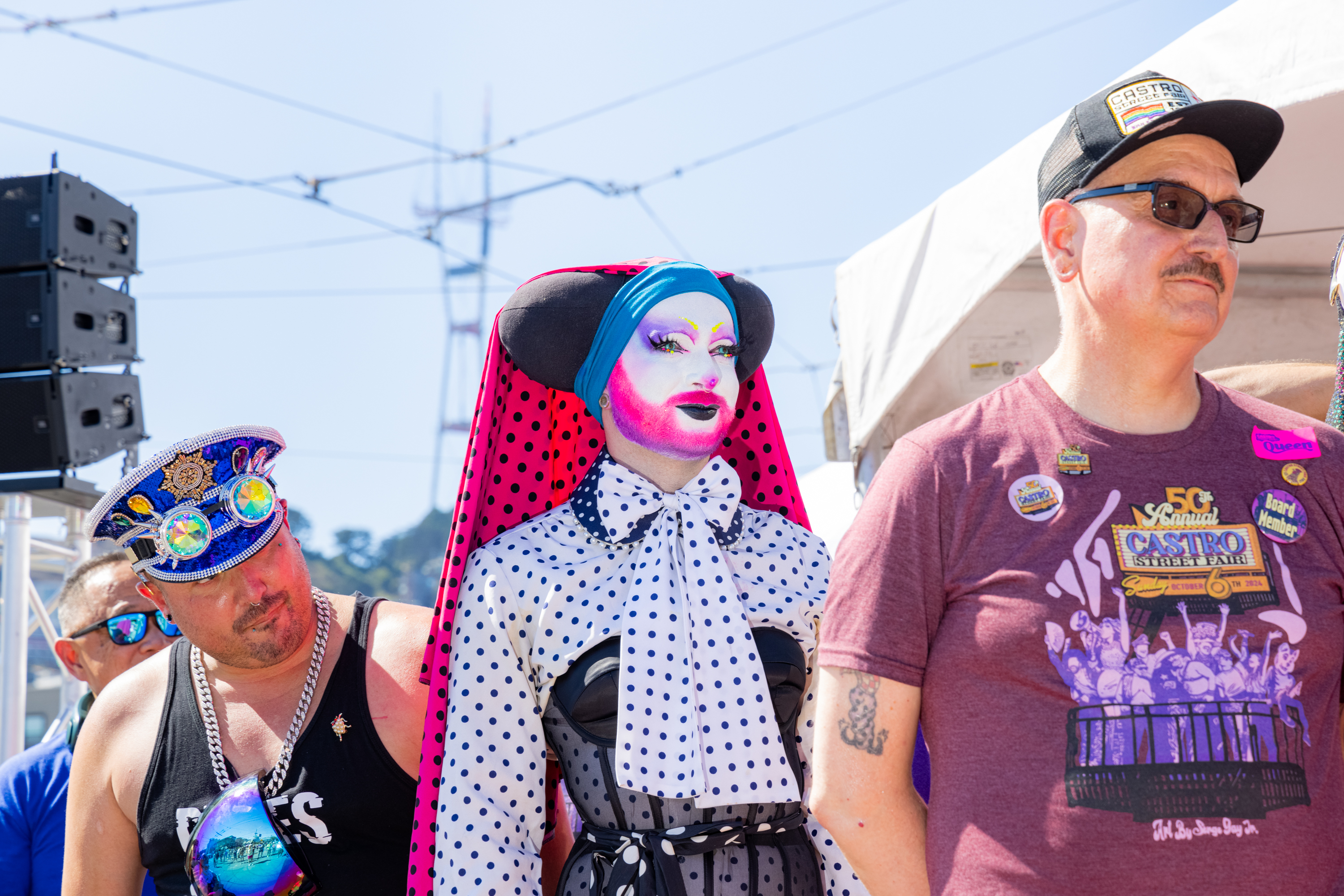 Three individuals in vibrant attire stand outdoors. One person is in colorful drag with a polka dot outfit. Another sports a bedazzled cap, and the third wears a festival shirt.