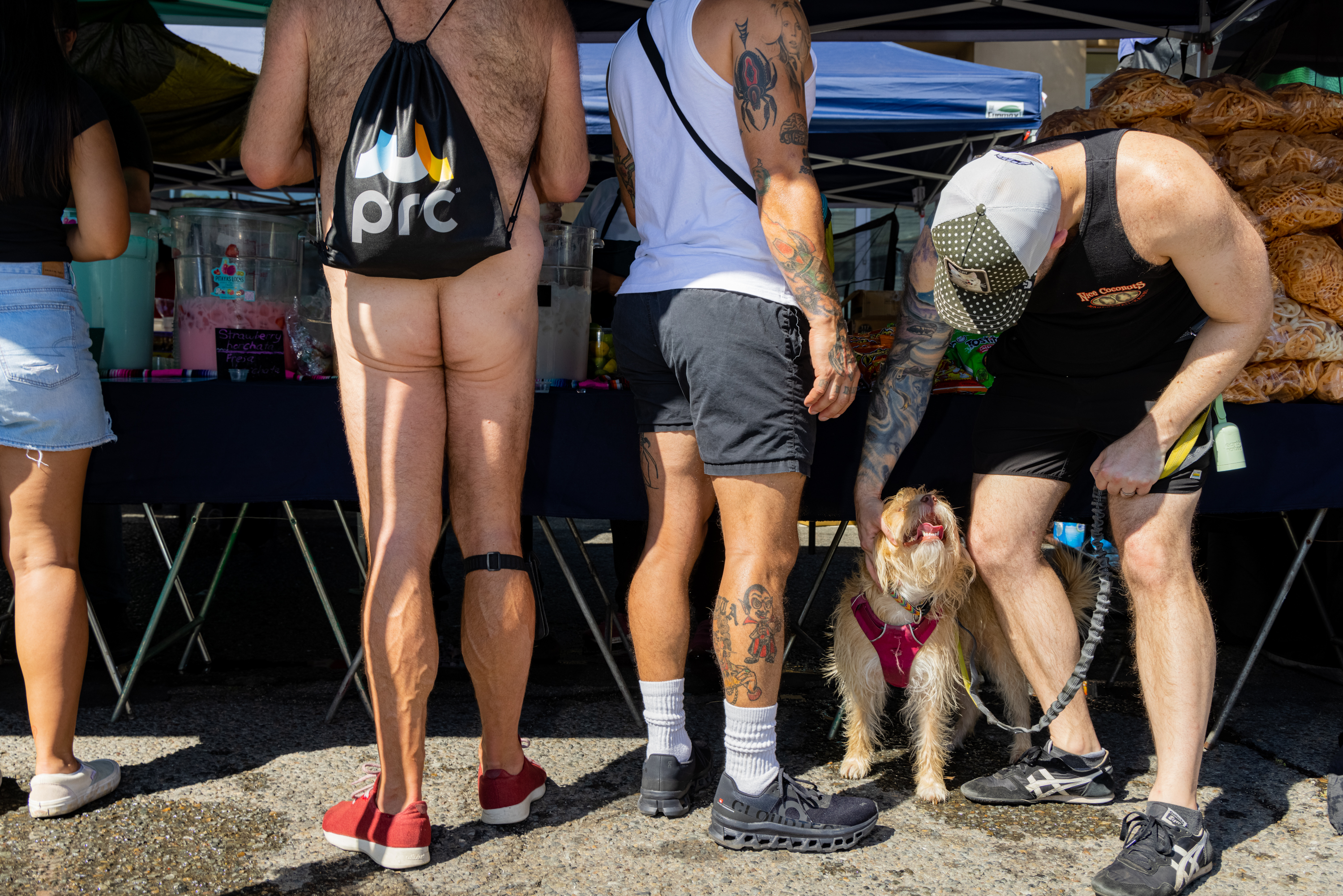 Several people stand at a market stall; one is naked except for a backpack. Another person pets a dog wearing a pink harness. Bags of snacks are visible.