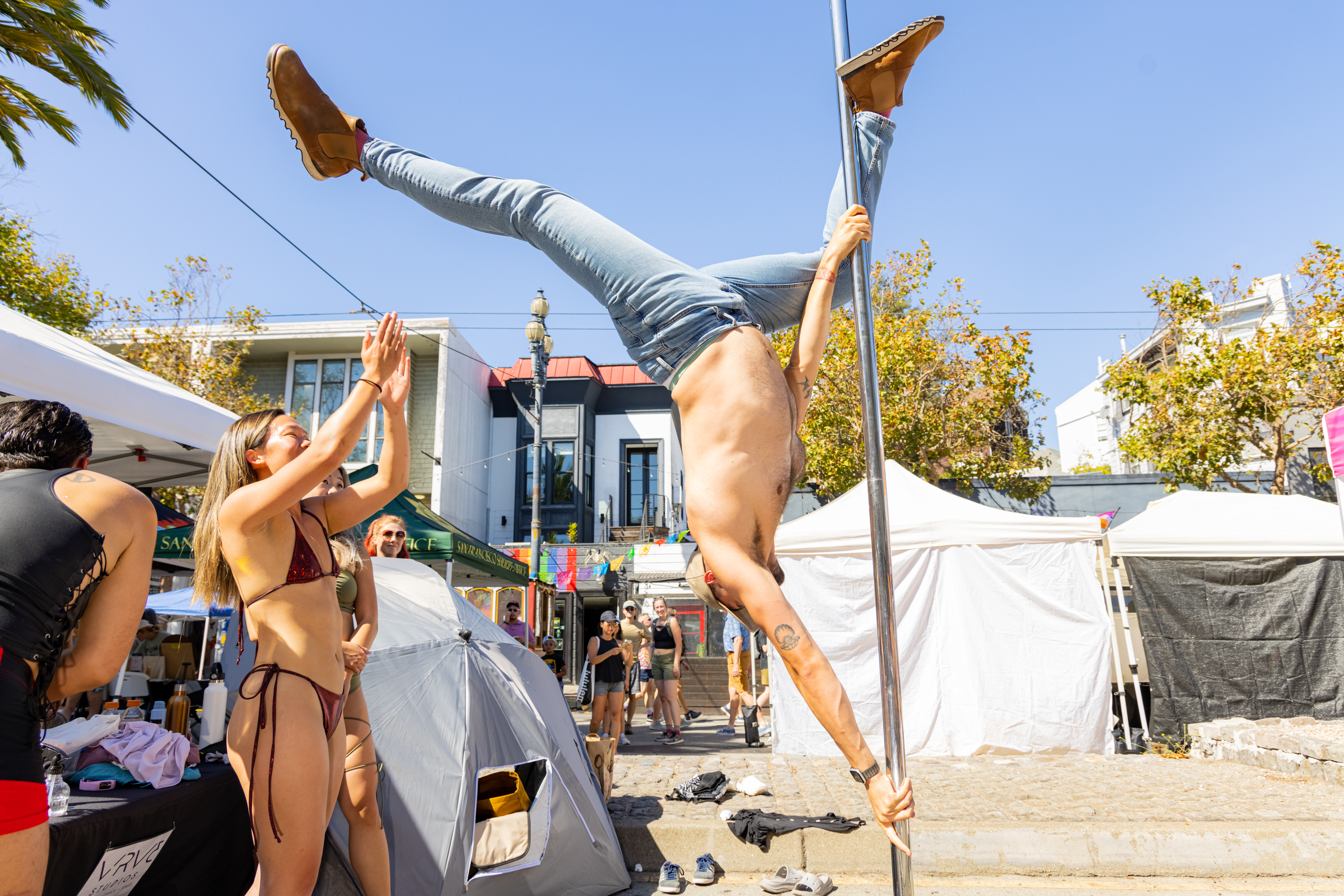 A man performs an acrobatic pole move, gripping it upside down, while a woman in a swimsuit applauds nearby. Tents and people fill the sunny street scene.