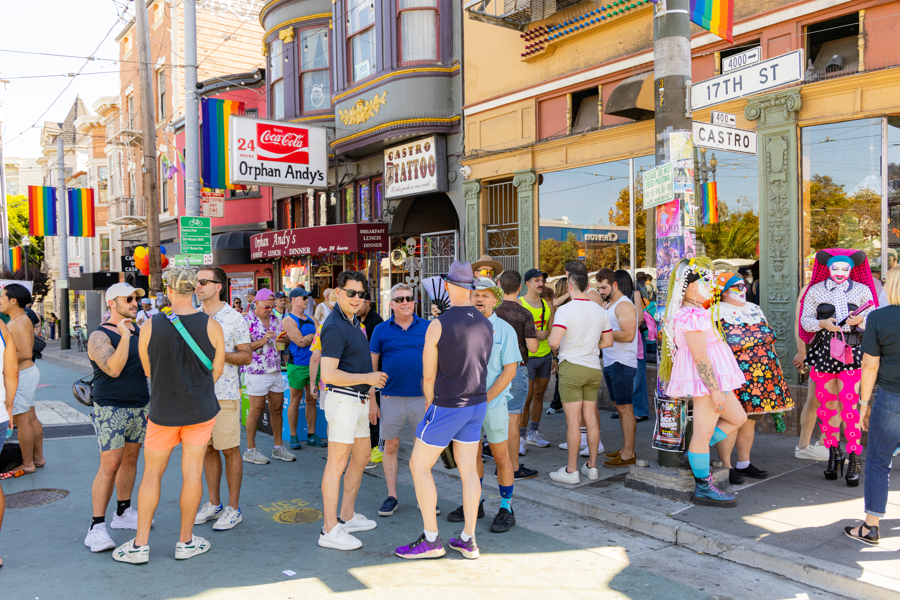 A lively group of people socializing on a colorful street corner with rainbow flags, vibrant outfits, and the sign for Castro Tattoo visible.