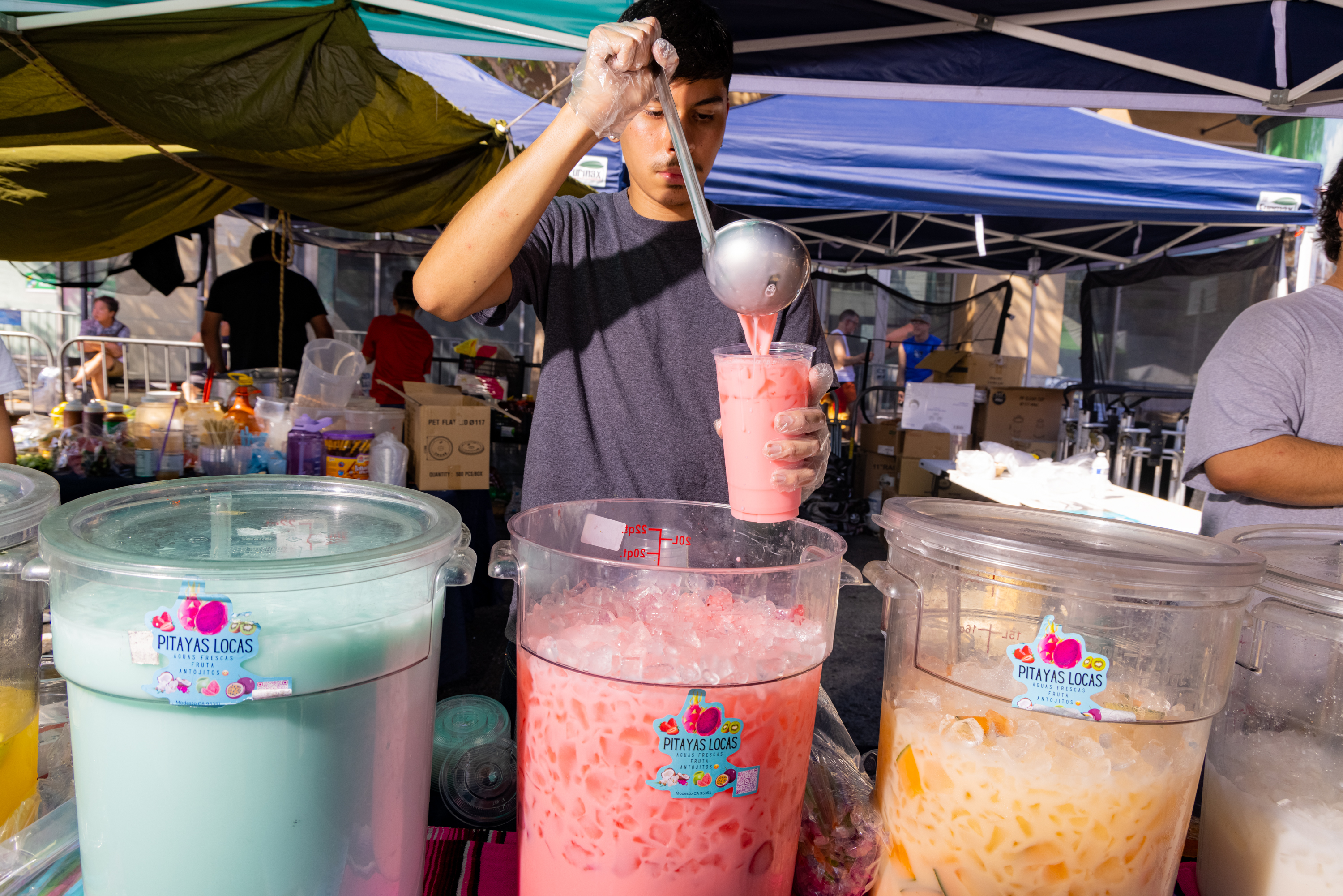 A person is ladling pink juice from a large container into a cup at an outdoor market stand. Multiple colorful drink containers surround them under a canopy.