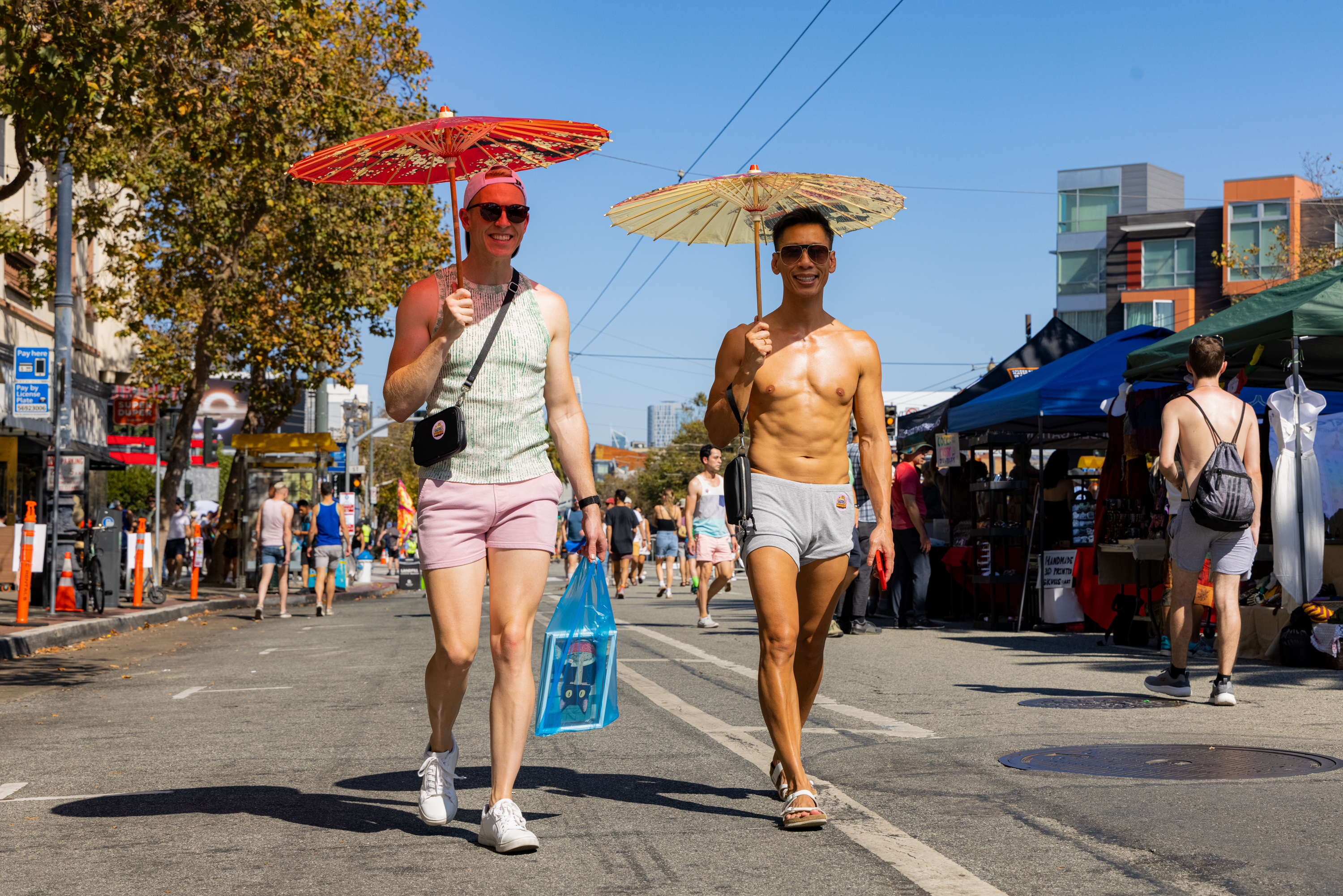 Two people walk side by side on a street, holding parasols. One is wearing a tank top and shorts, the other is shirtless in shorts. The street is lined with market stalls.