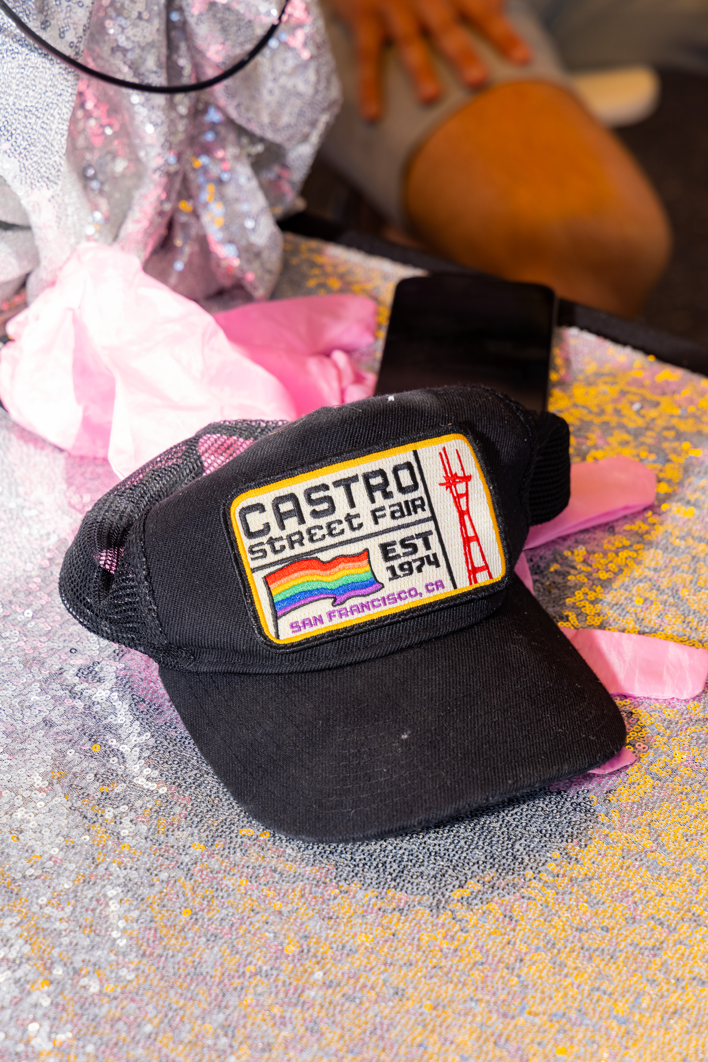 A black cap with a &quot;Castro Street Fair&quot; patch featuring a rainbow and red tower lies on a sparkly surface with pink fabric nearby.