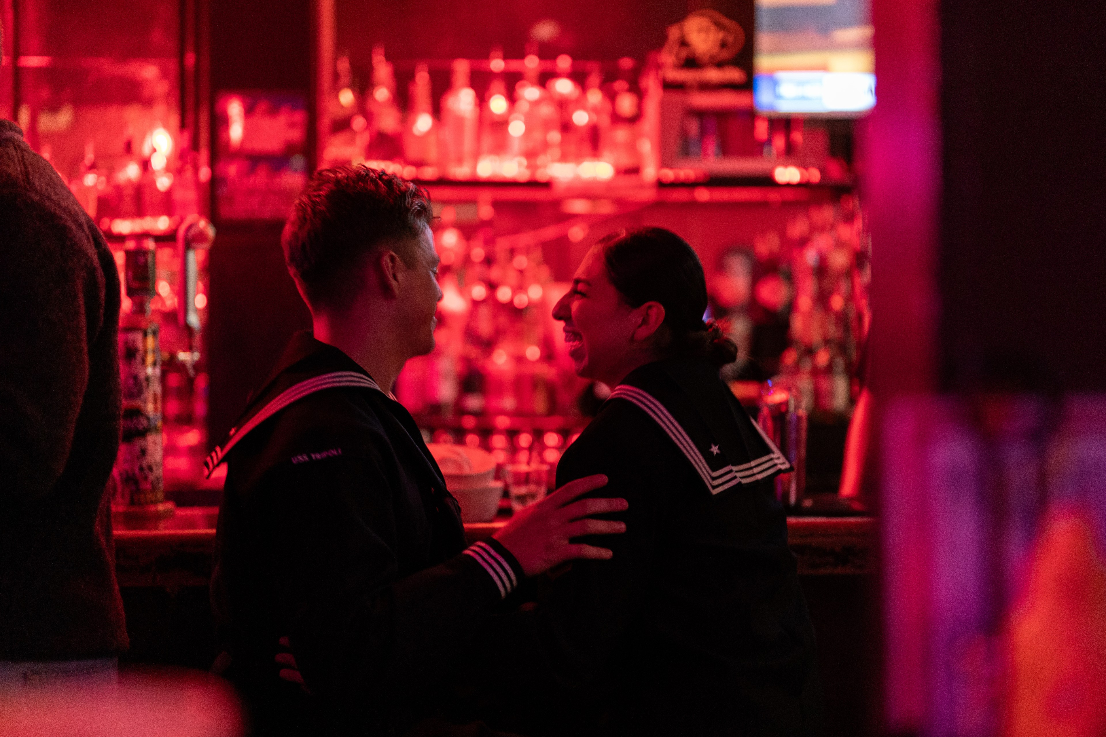 Two people in sailor uniforms are seated at a bar, engaged in conversation. The atmosphere is dimly lit with a red hue, and bottles line the shelves behind them.