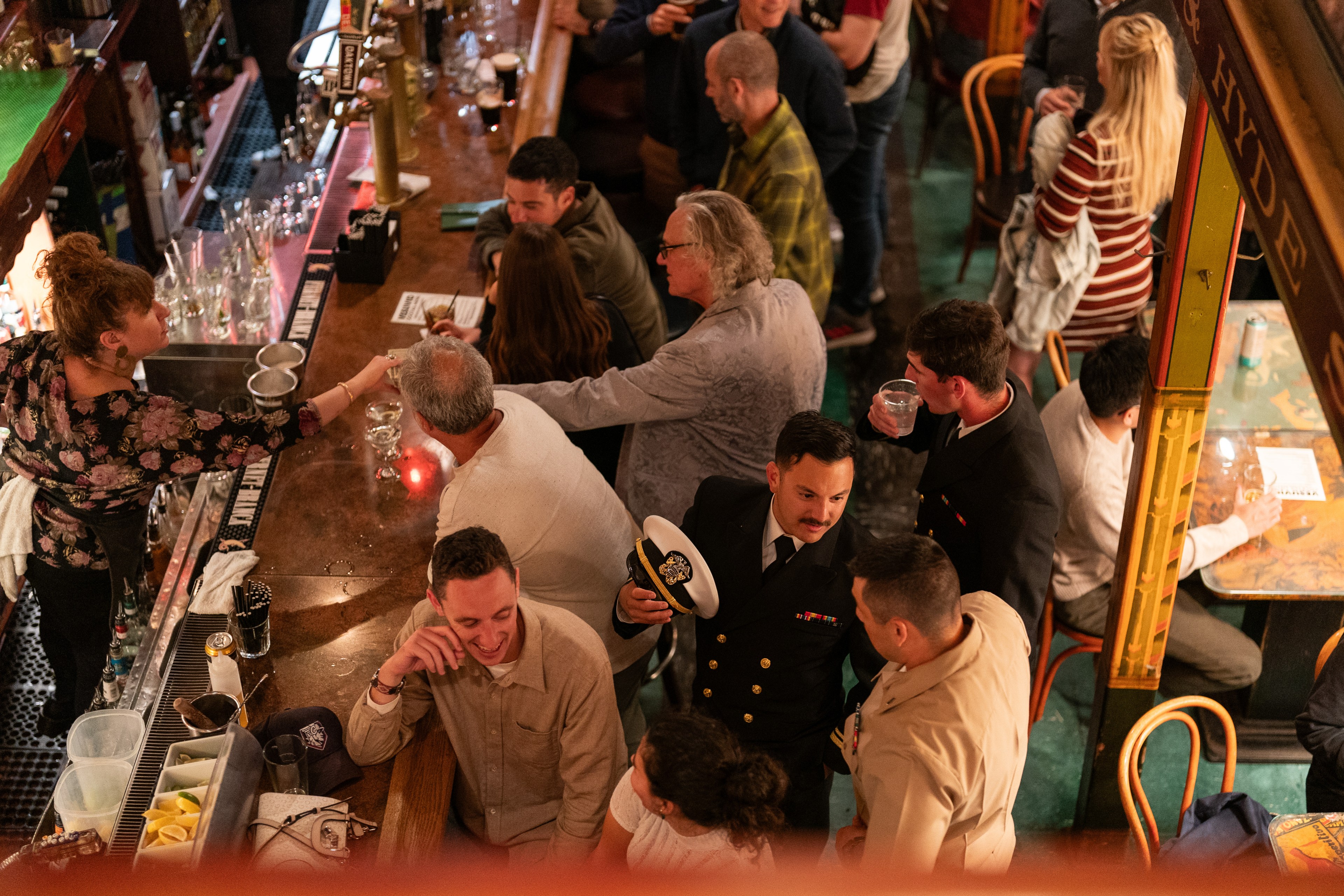 A busy bar scene features people socializing; a bartender serves drinks to patrons seated along a wooden counter. A man in a uniform holds a cap, talking to others.