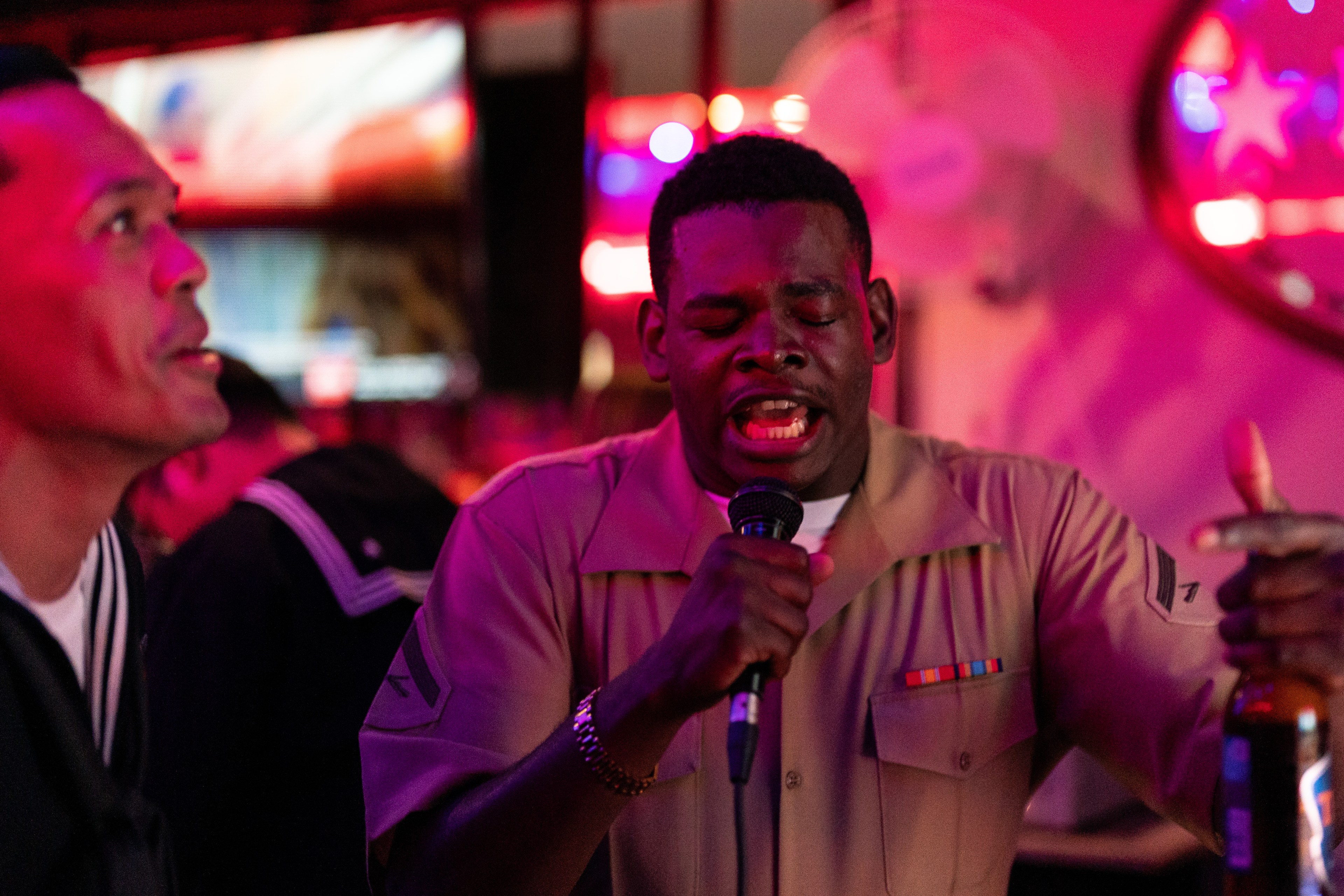 Two men are singing karaoke in a dimly lit bar with red lighting. One man passionately holds a microphone, while the other listens, dressed in uniform.