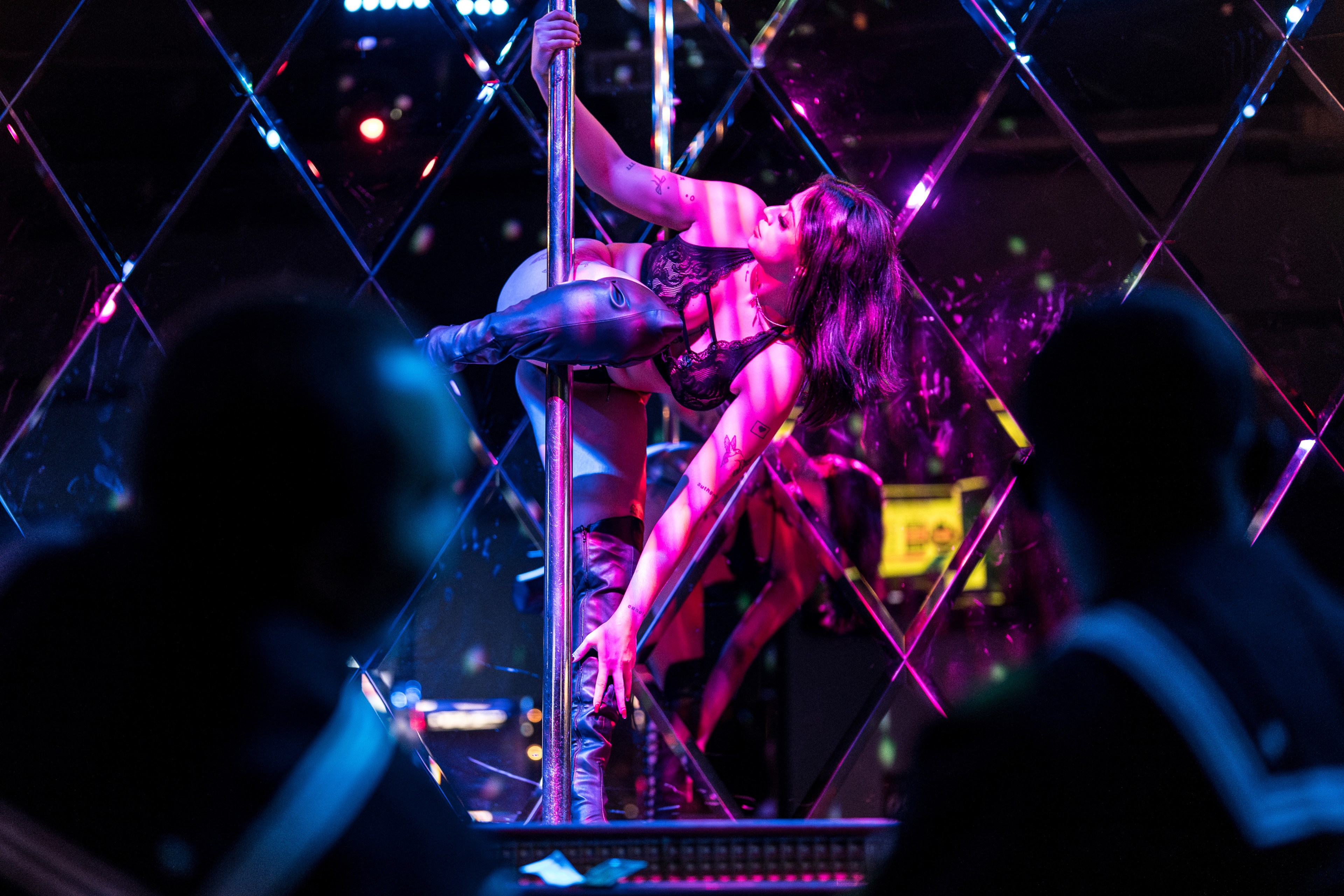 A person performs pole dancing in a dimly lit, colorful room with mirrored walls. They are wearing a black outfit, and the lighting creates a vibrant, stage-like atmosphere.