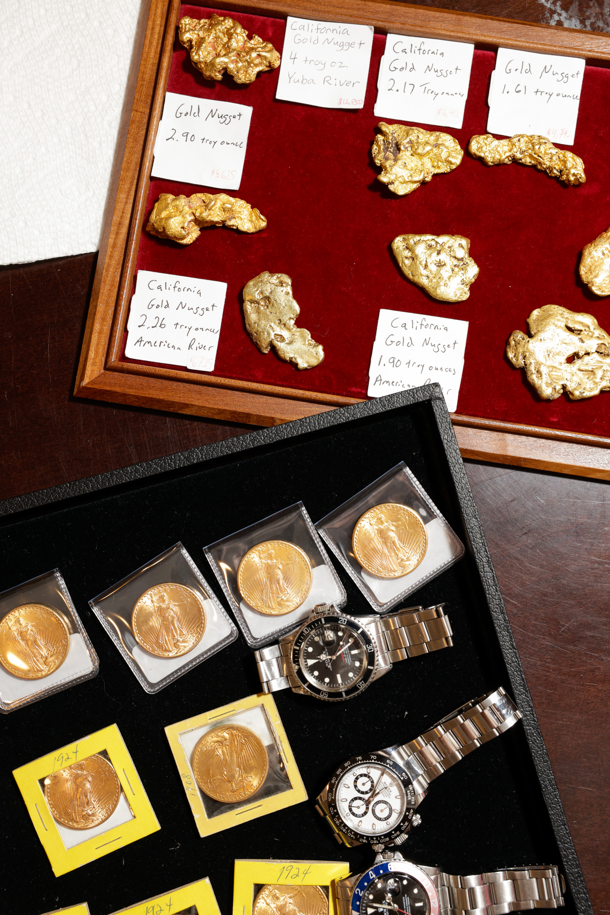 The image shows a display of gold nuggets labeled with weights, gold coins in protective cases, and three luxury watches with metal bands arranged neatly.