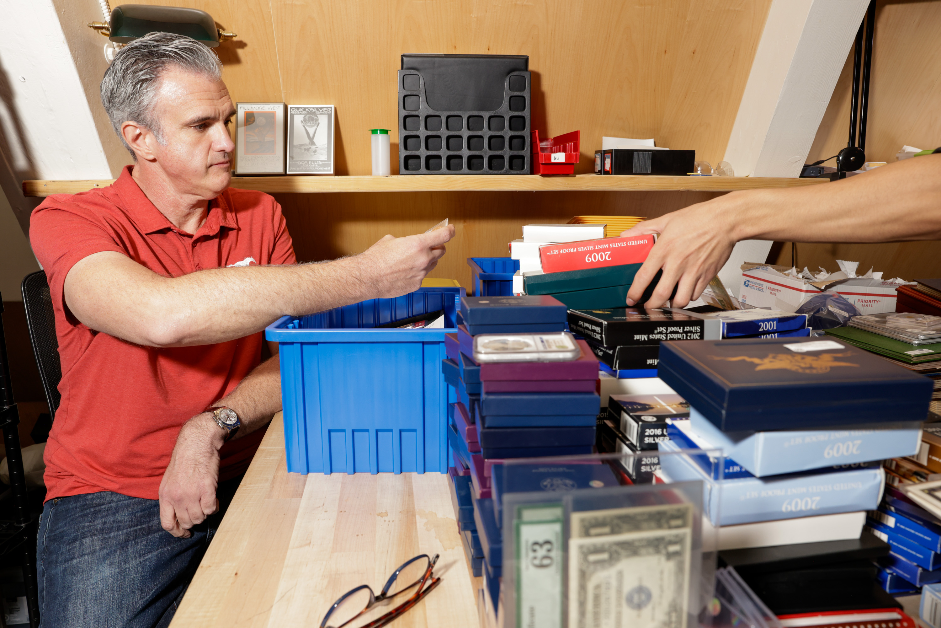 A man in a red shirt examines items on a table filled with various boxes and containers, including a stack of U.S. Mint silver proof sets and currency.