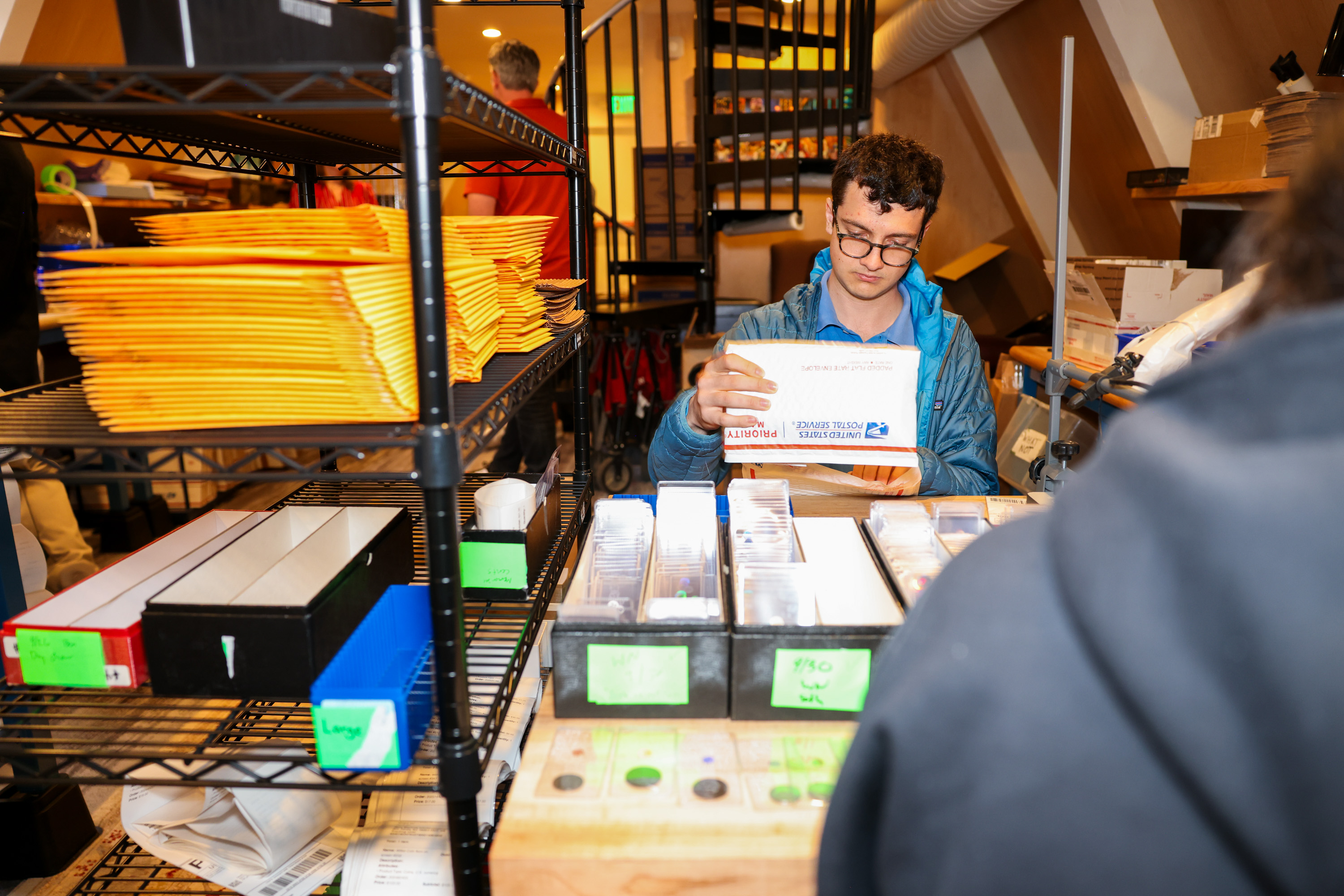 A person sorts packages at a table in a cluttered room with shelves full of yellow envelopes and various supplies.