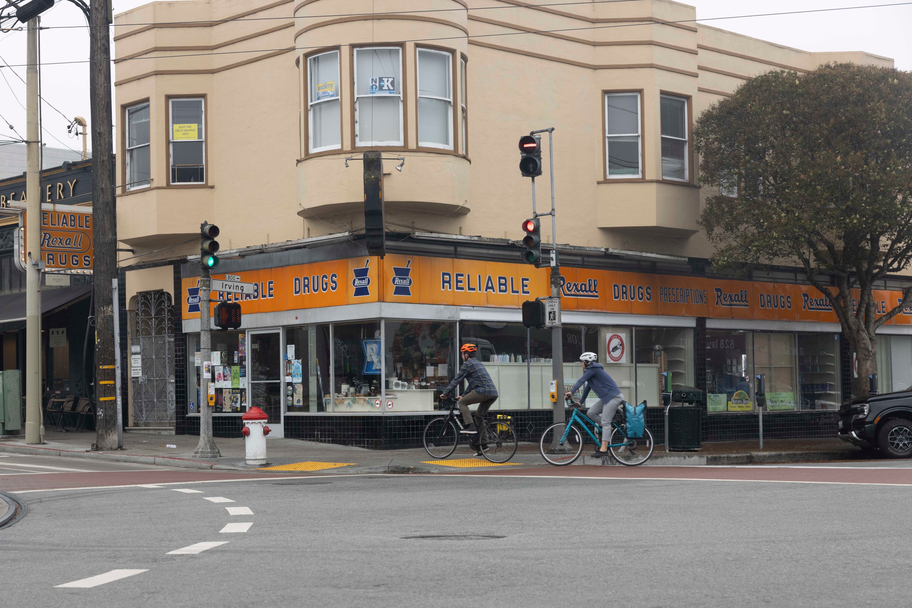 Two cyclists ride past a corner drugstore with an orange and blue &quot;Reliable Drugs&quot; sign. The building is beige with large windows above the storefront.