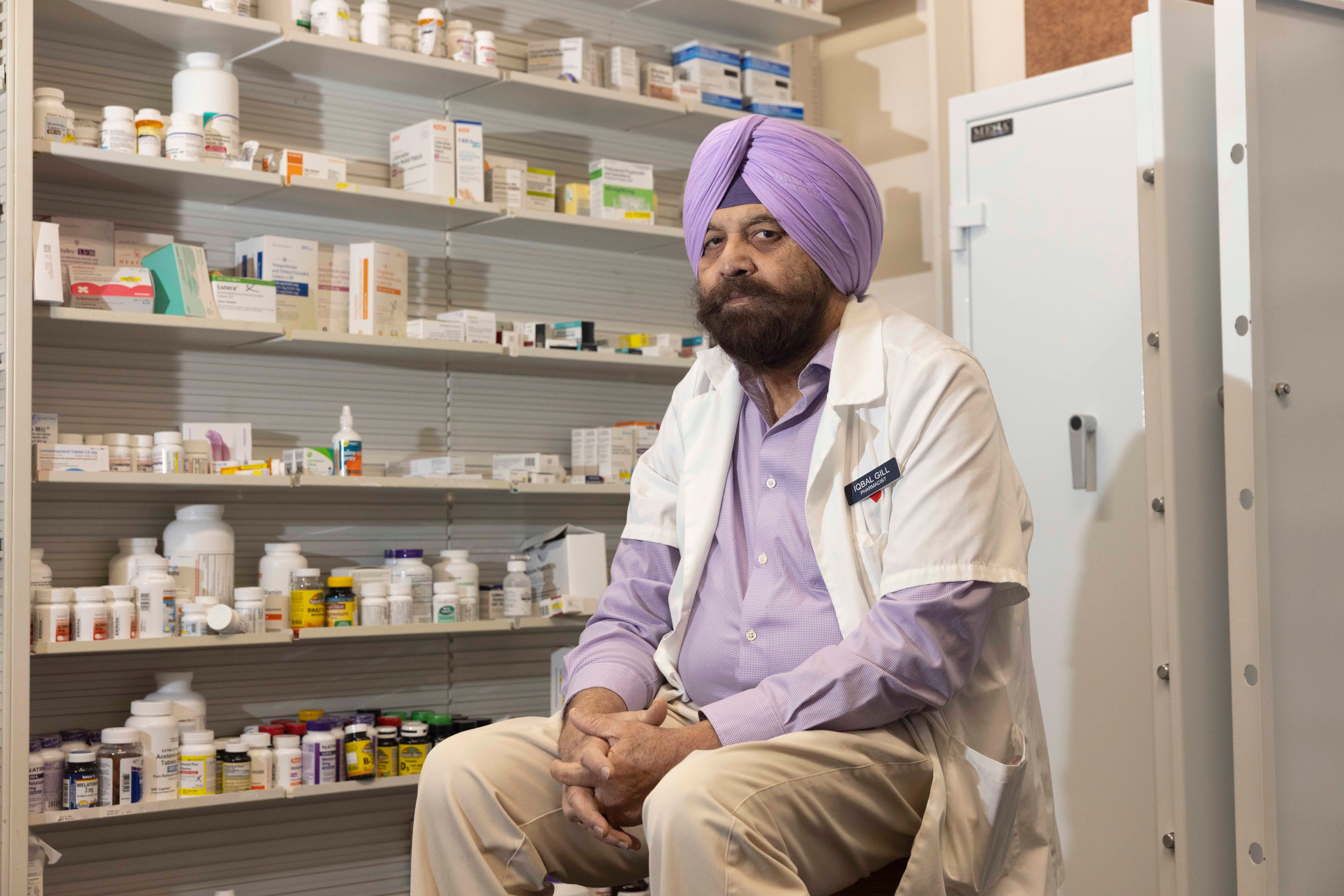 A man in a purple turban and white lab coat sits in a pharmacy, surrounded by shelves filled with various medicine bottles and boxes.