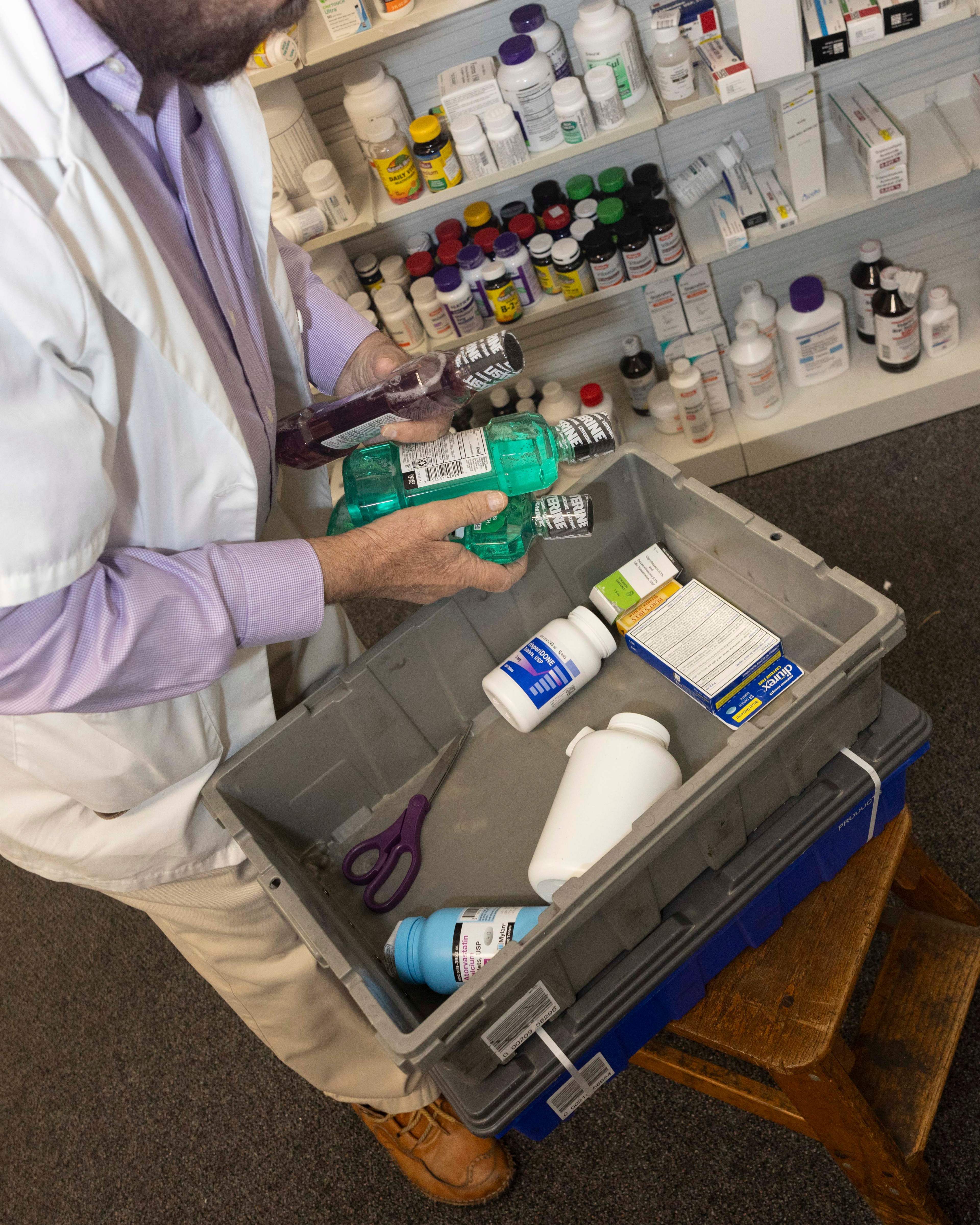 A person in a white lab coat is holding bottles of mouthwash above a bin with medicine bottles, scissors, and boxes. Shelves of more medicine are in the background.