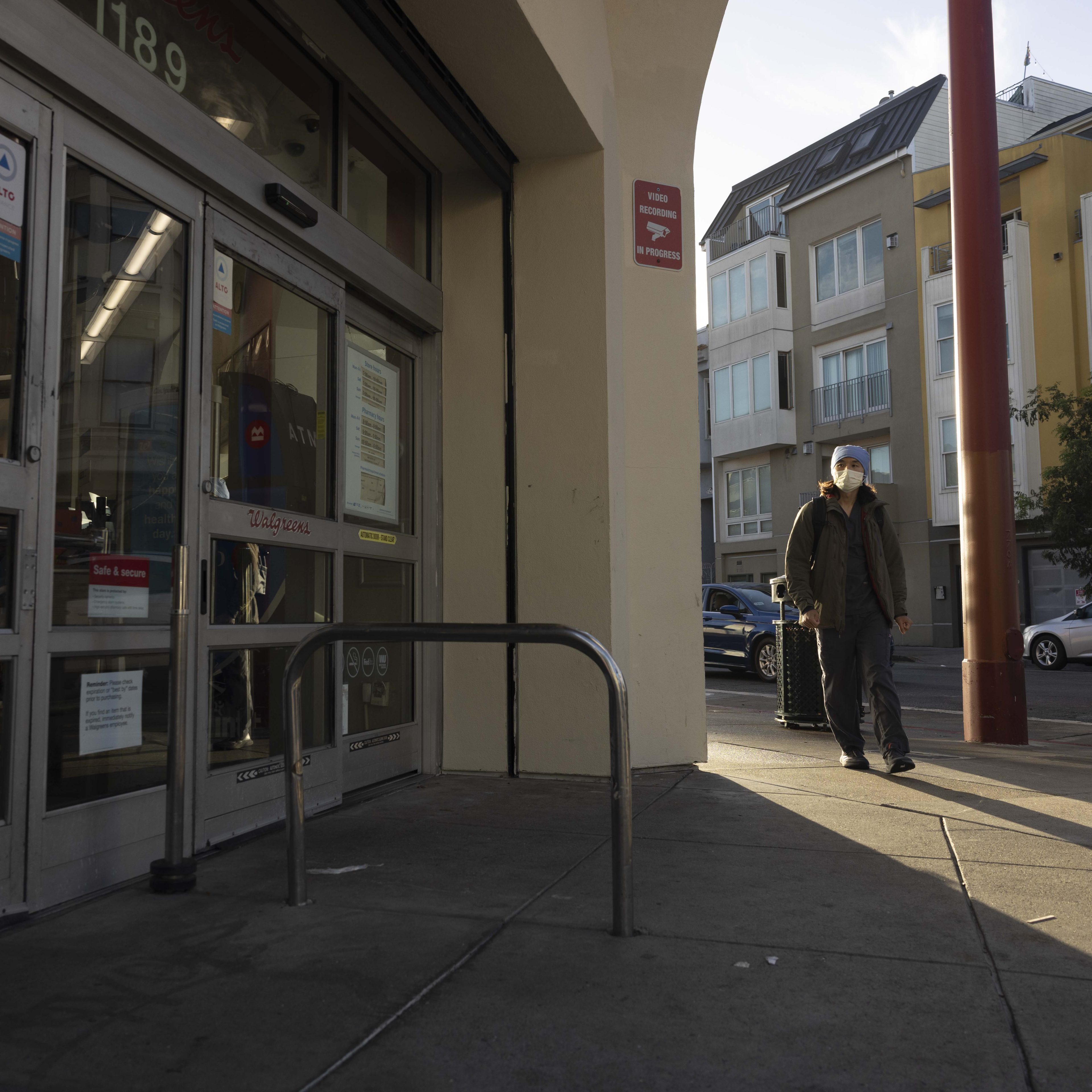 A person wearing a mask walks by a Walgreens entrance with glass doors on a city street. Sunlight casts long shadows, and nearby buildings are visible.