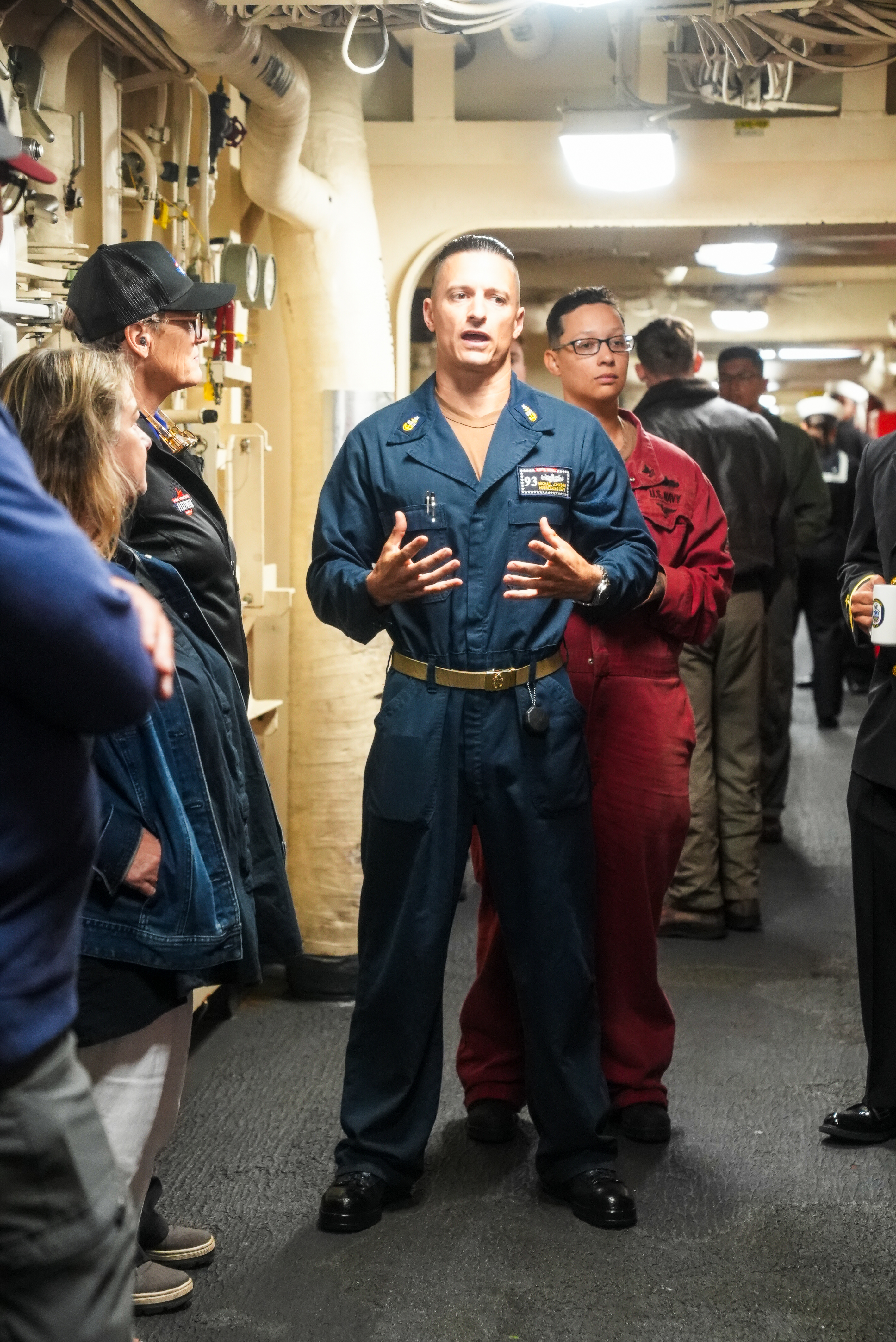 A man in a dark blue jumpsuit is speaking to a group in a narrow corridor, with pipes overhead. Others in uniforms are listening attentively.