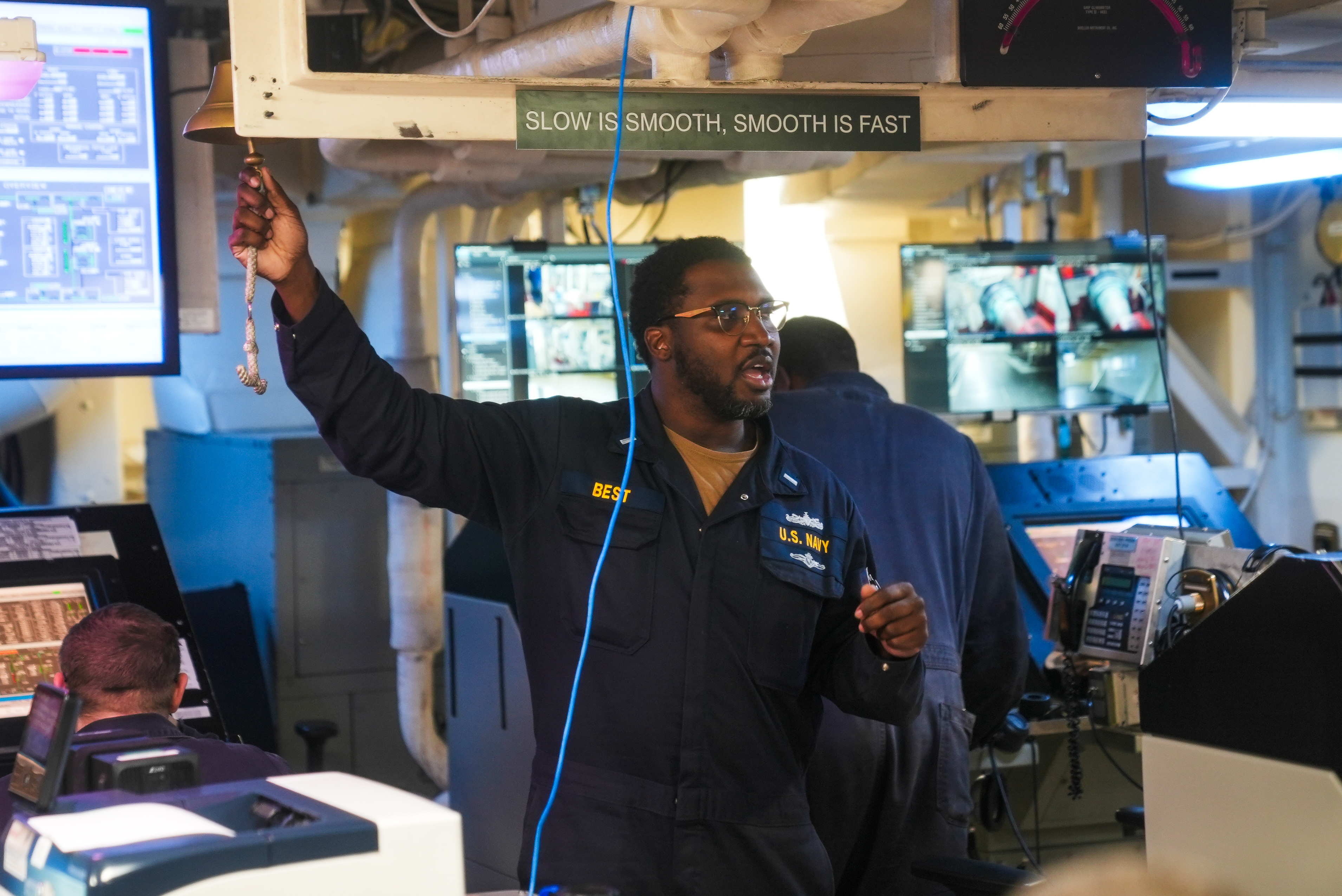 A person in a U.S. Navy uniform stands inside a control room, ringing a bell. Monitors are visible, and a sign above reads, &quot;Slow is smooth, smooth is fast.&quot;