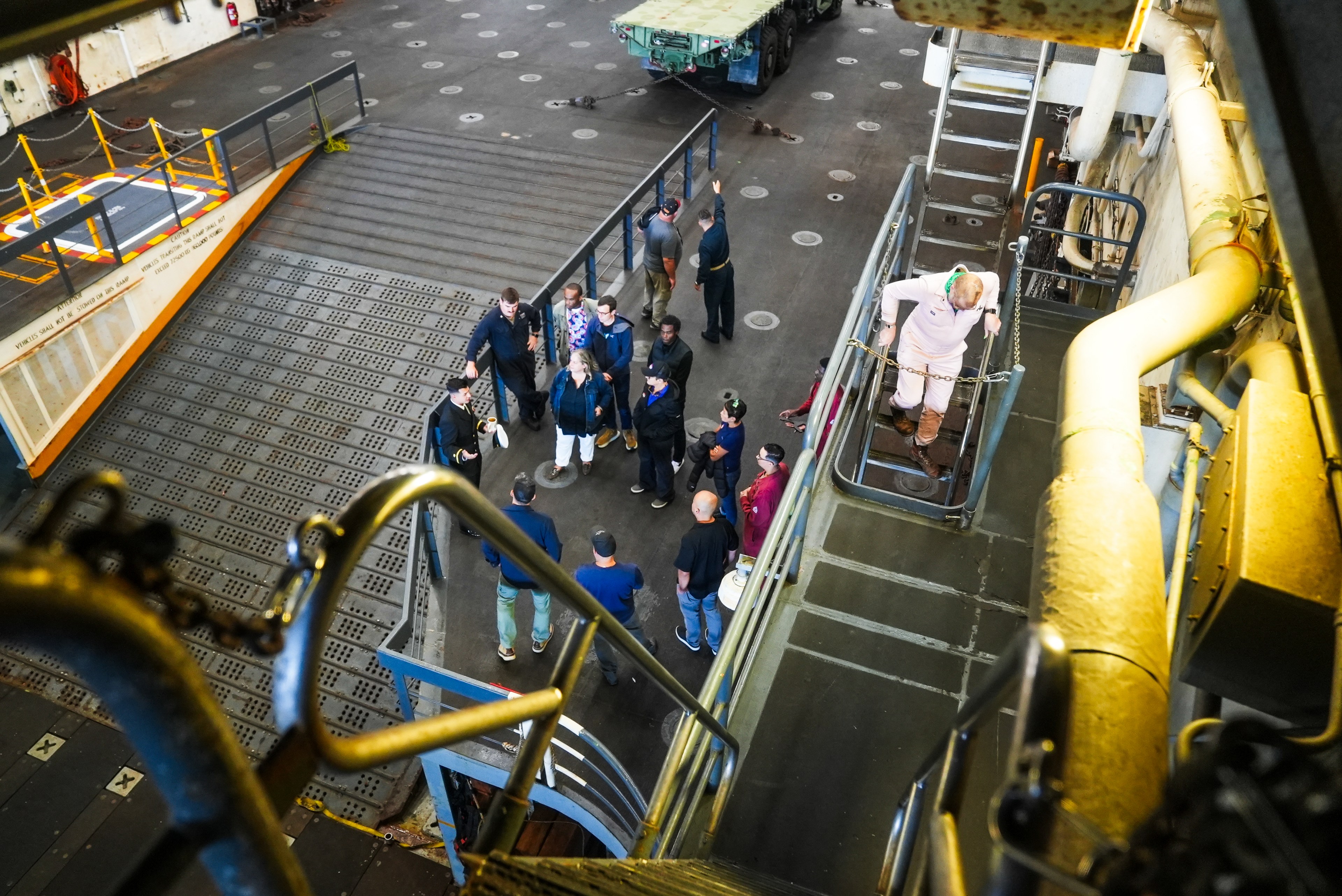 A group of people stands on a lower level of a ship's interior, near a ramp and a parked vehicle. One person is on a staircase leading to the upper level.