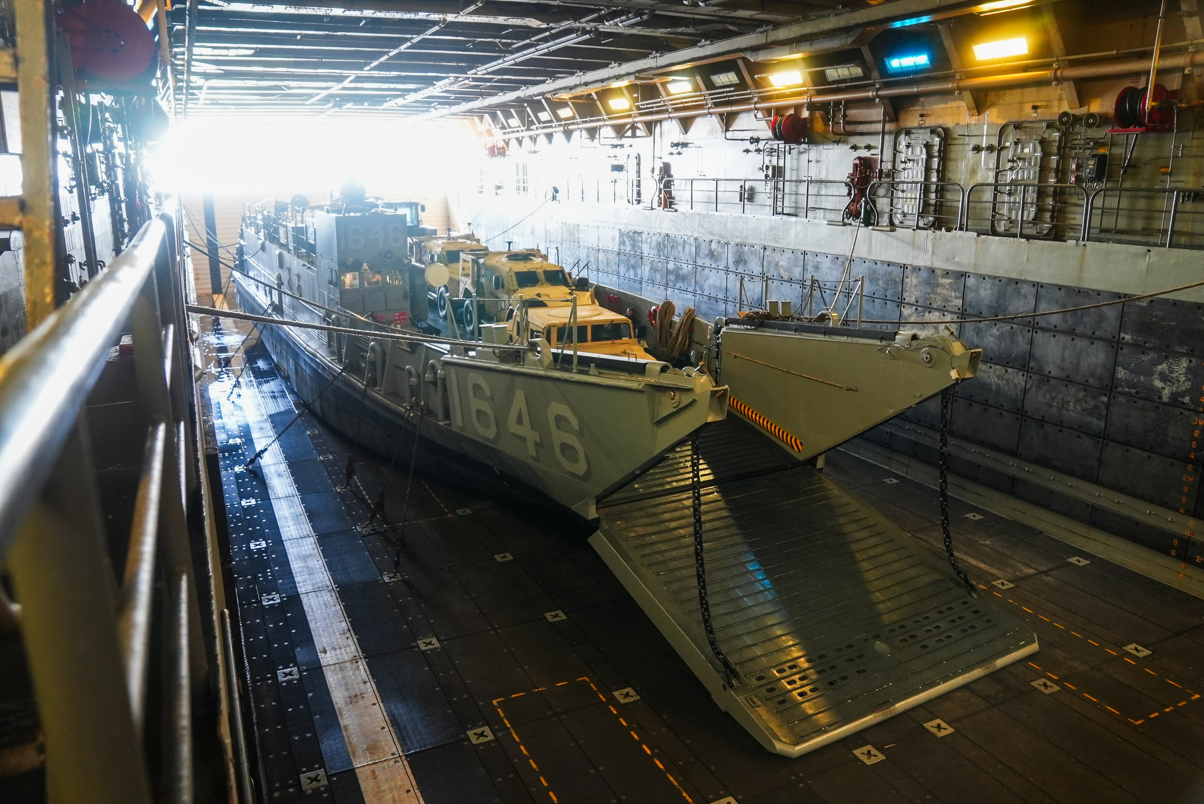 A military landing craft with the number 1646 is in the bay of a larger ship. It has a lowered ramp, and several military vehicles are aboard.