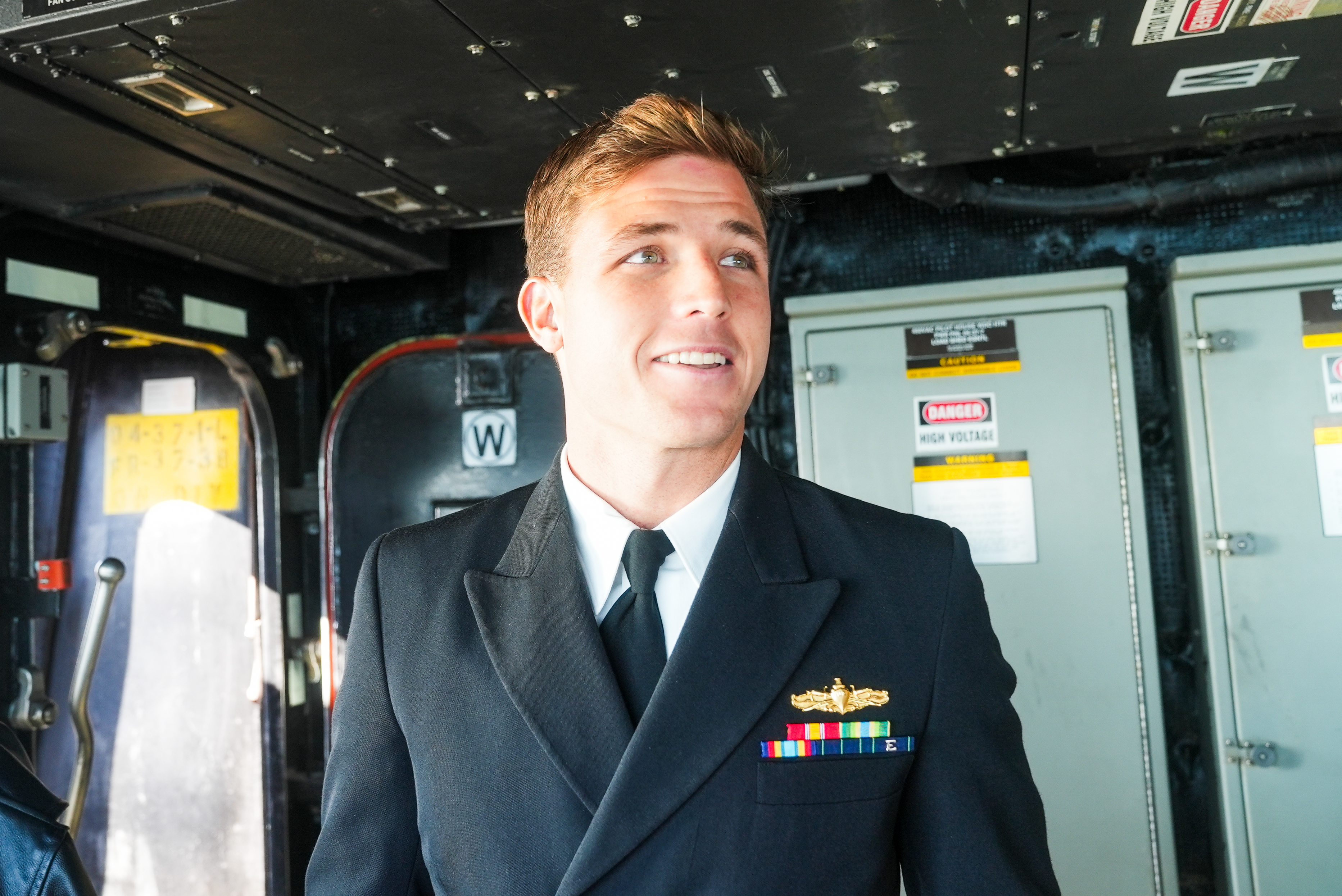 A man in a military uniform with colorful ribbons and a golden badge stands inside a control room, smiling. Electrical panels labeled &quot;HIGH VOLTAGE&quot; are visible.