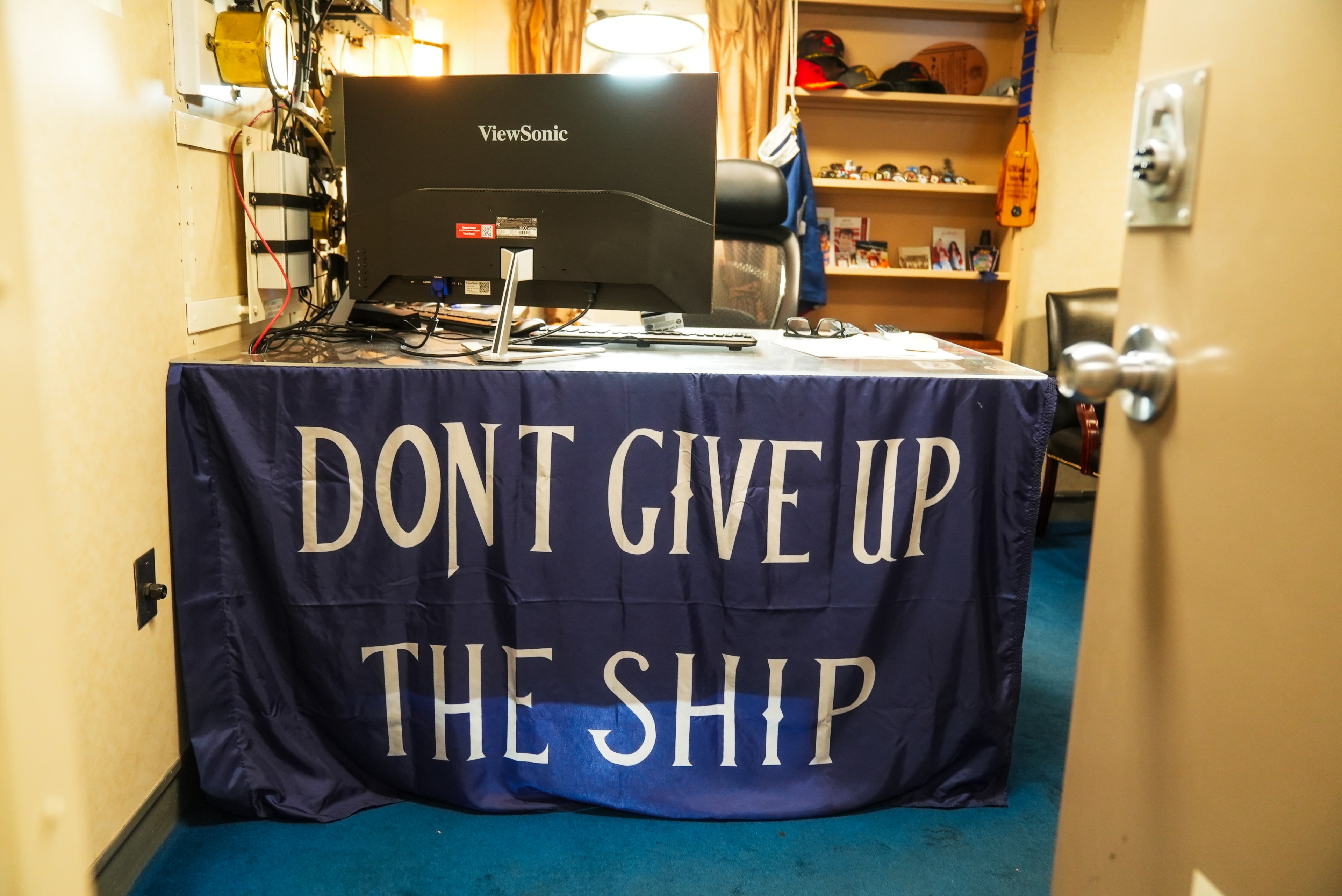 The image shows a desk with a ViewSonic monitor. A blue flag draped over the desk reads &quot;DON'T GIVE UP THE SHIP.&quot; A shelf with hats and photos is in the background.