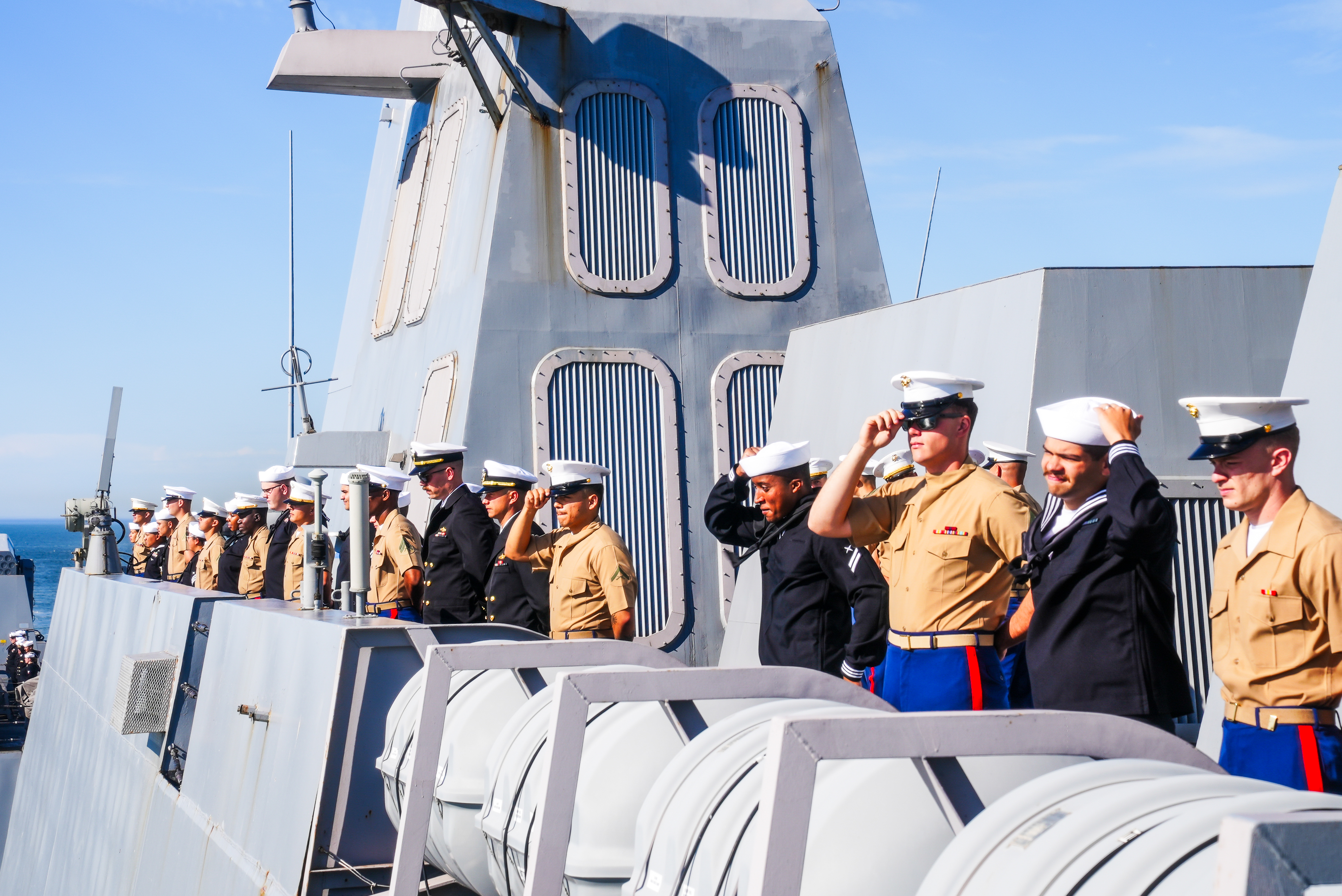 Sailors and marines in uniform stand on the deck of a naval ship, some saluting, against a backdrop of the ship's structure and a clear blue sky.