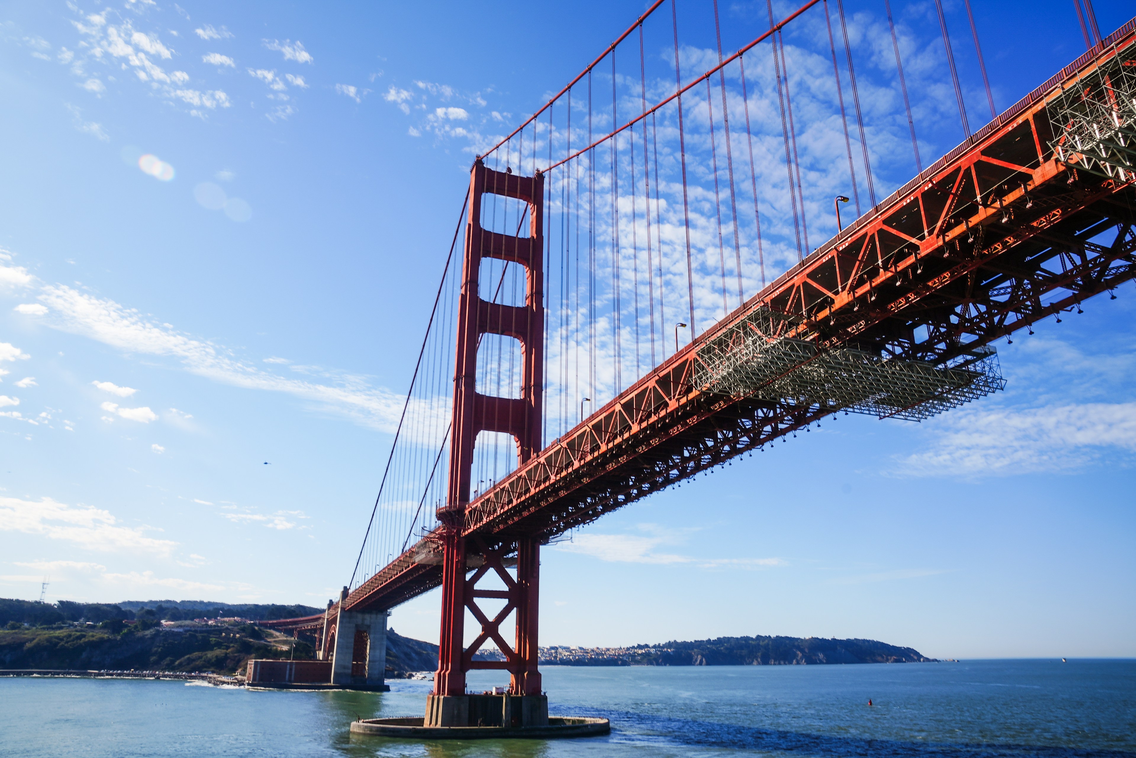 A red suspension bridge stretches across a body of water under a clear blue sky, with distant hills and scattered clouds in the background.