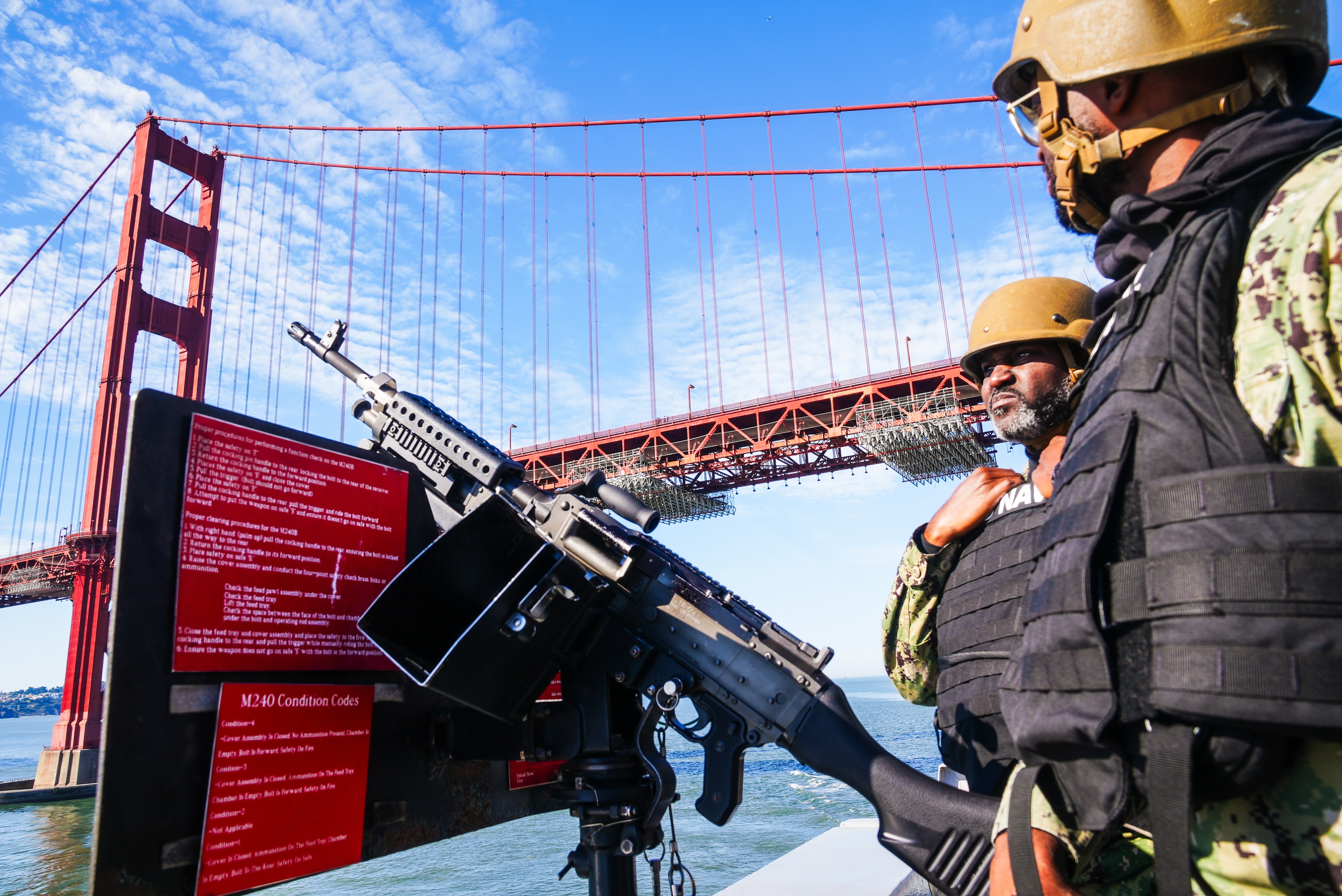 Two people in tactical gear stand next to a mounted machine gun, with the Golden Gate Bridge visible in the background under a blue sky.