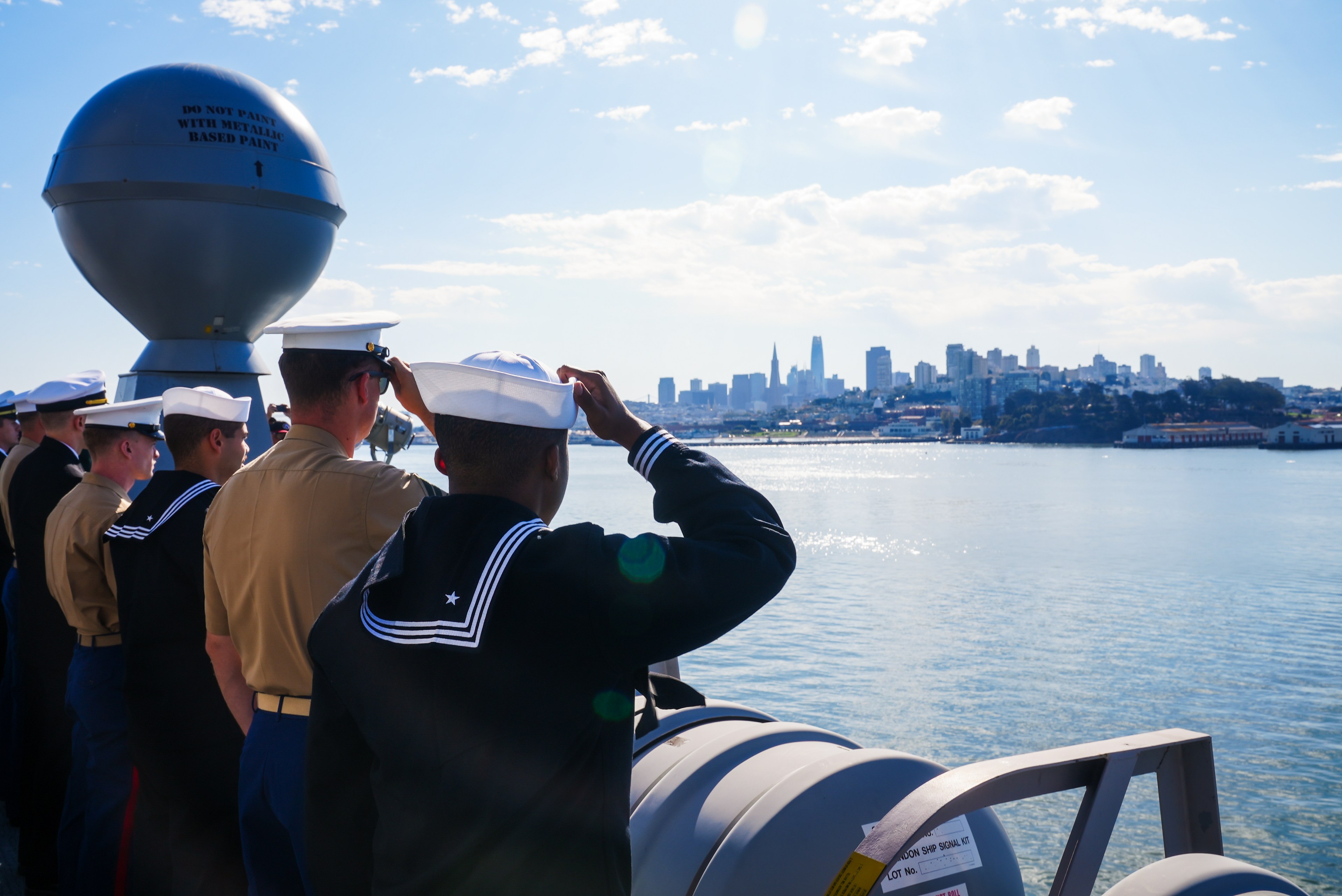 Sailors in uniform stand on a ship, gazing at a distant city skyline under a clear sky, with water surrounding them.