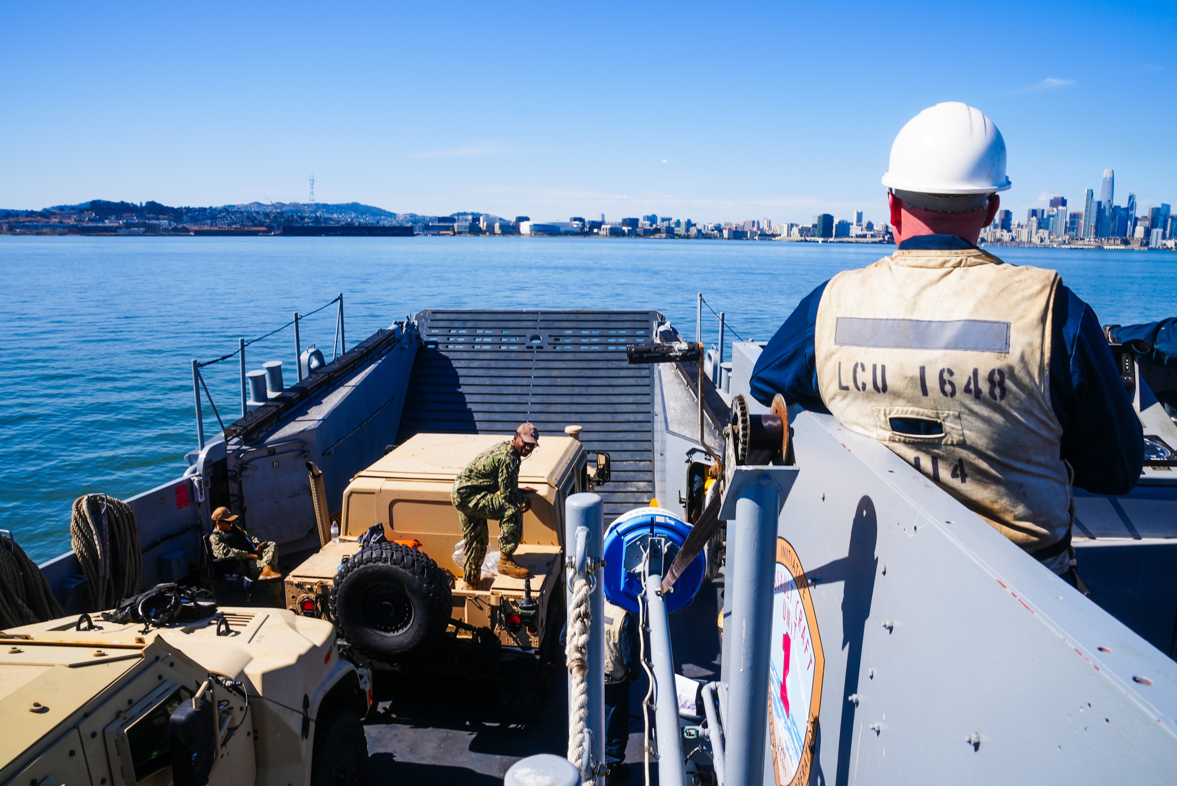 A person in a white helmet overlooks a landing craft with military vehicles. City skyline and blue water surround under clear blue skies.