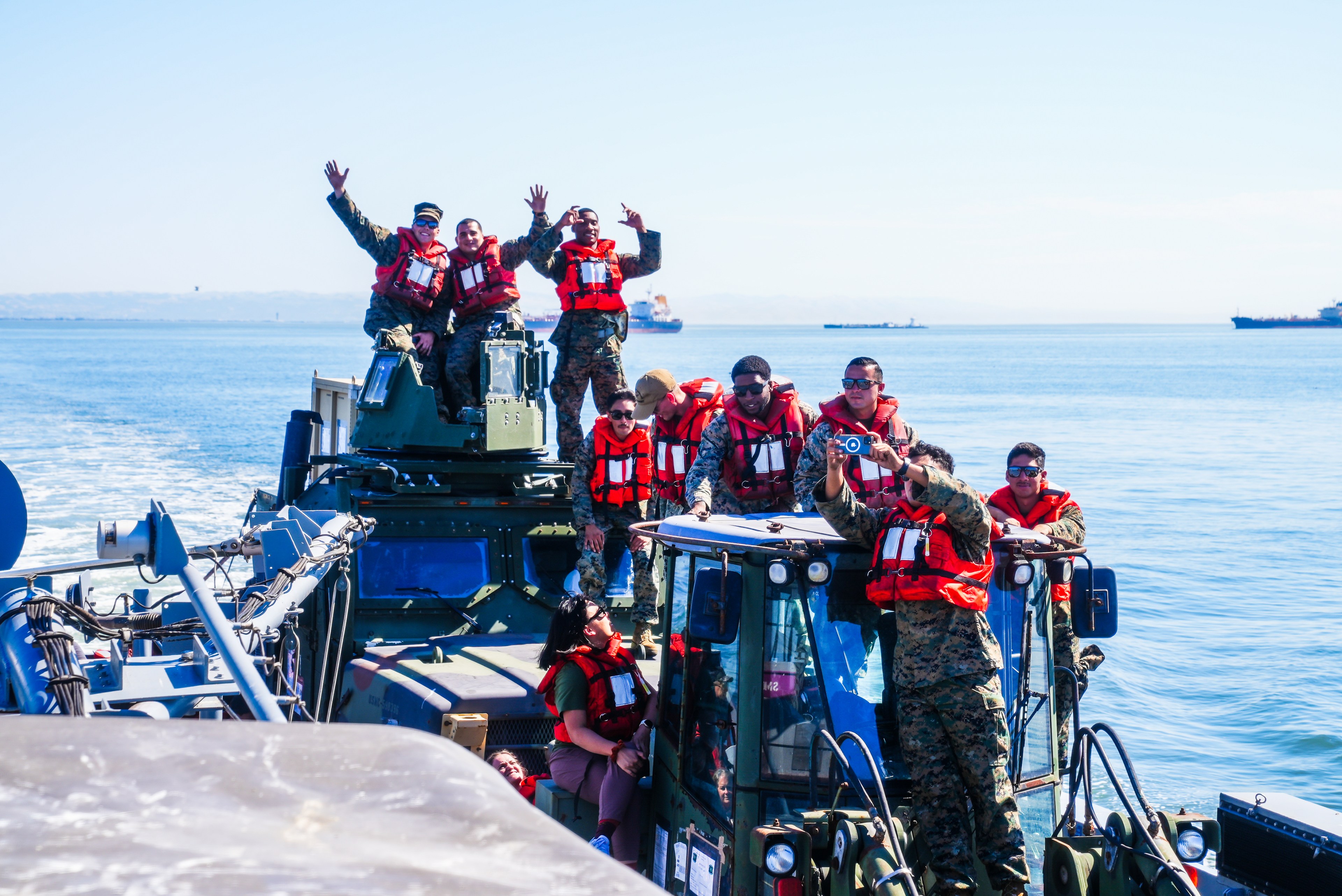 A group of people in military uniforms and red life jackets are on a boat, posing and smiling for a photo, with a clear blue sea in the background.