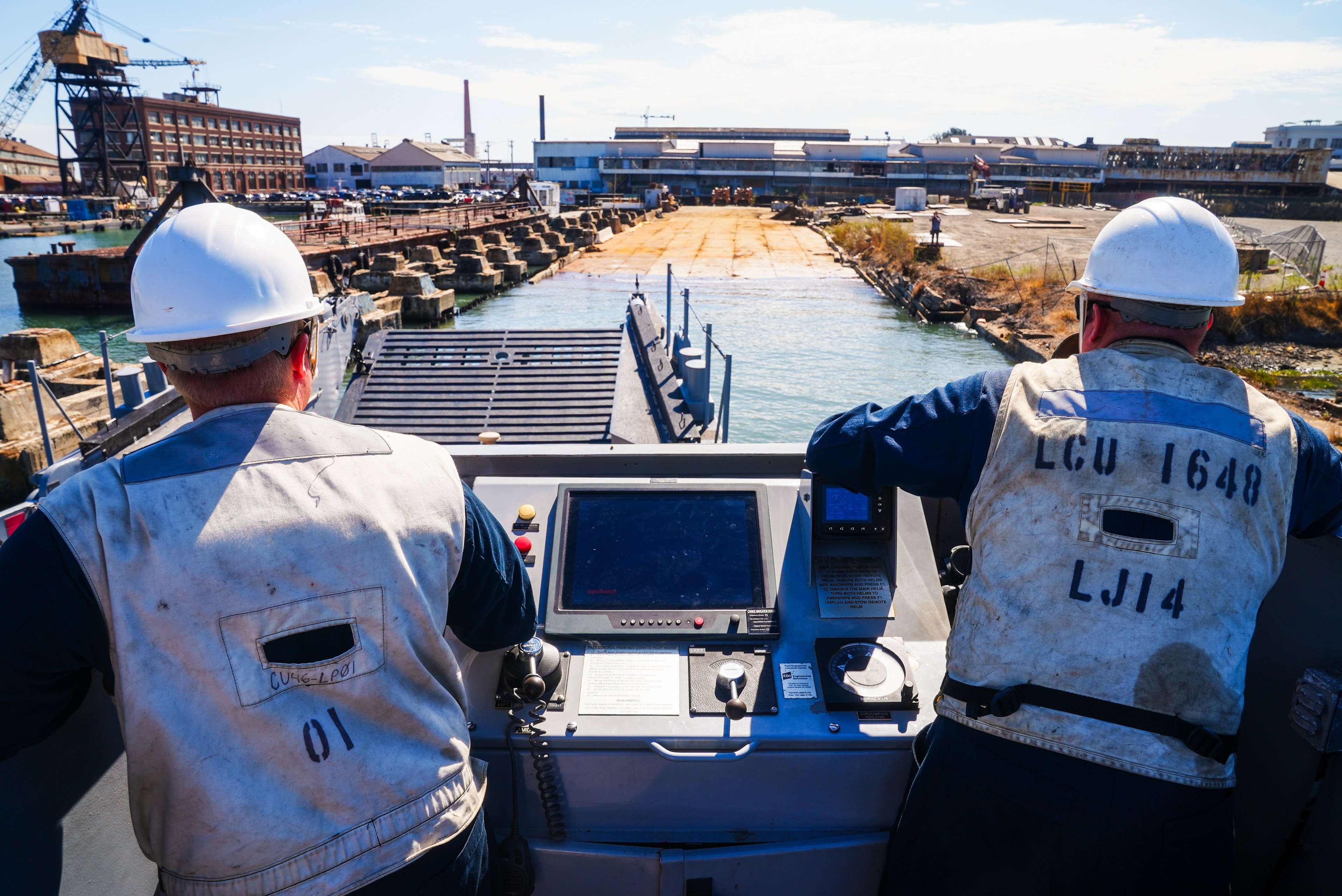 Two workers in hard hats and vests operate a control panel on a vessel near an industrial dock, with buildings, cranes, and water visible ahead.