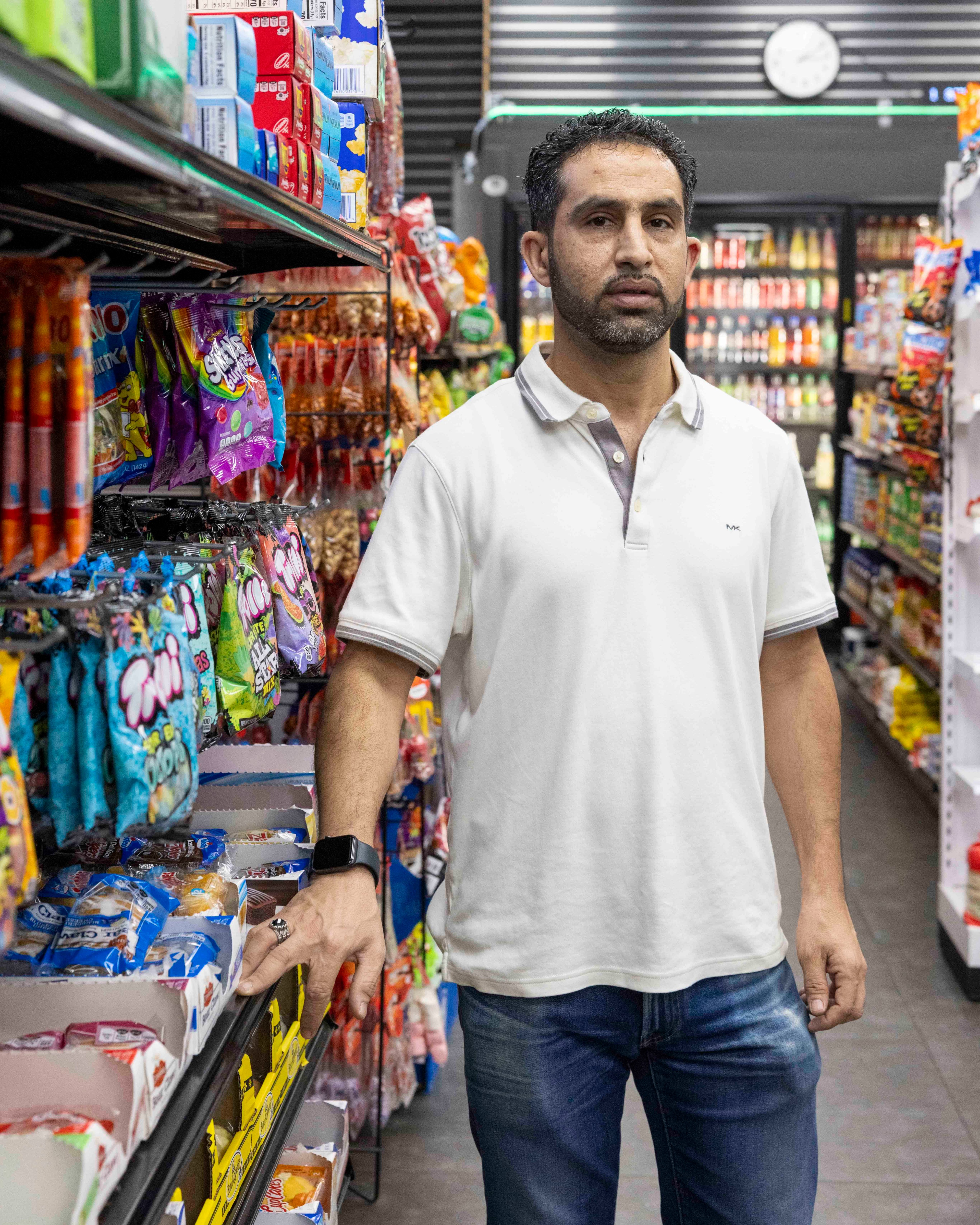 A man in a white polo shirt stands in a grocery aisle surrounded by colorful snack packages. He has a smartwatch on his wrist and a clock is visible behind him.