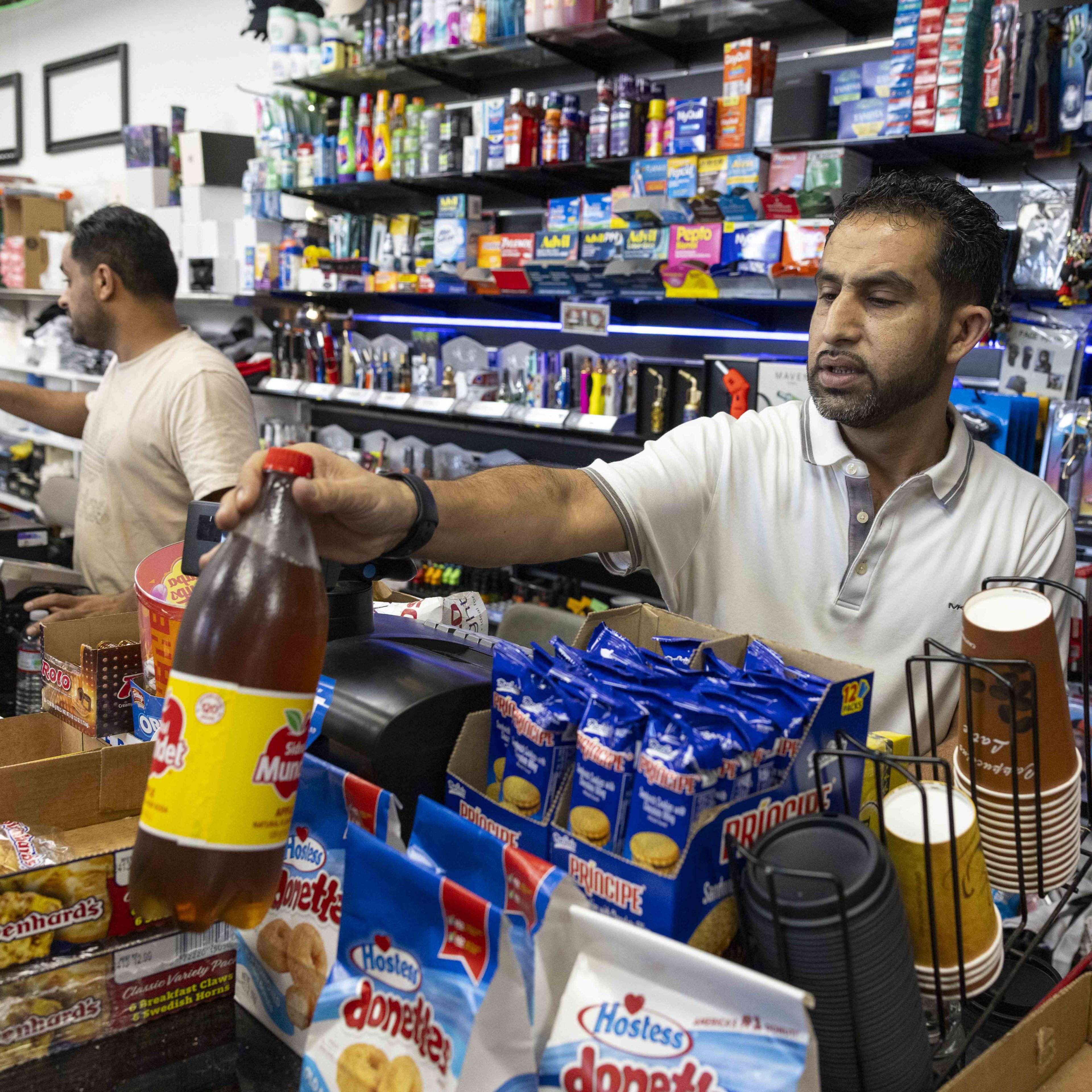 A man in a store is placing a large bottle of soda by the register. The counter is stocked with snacks, and shelves behind him display various products.