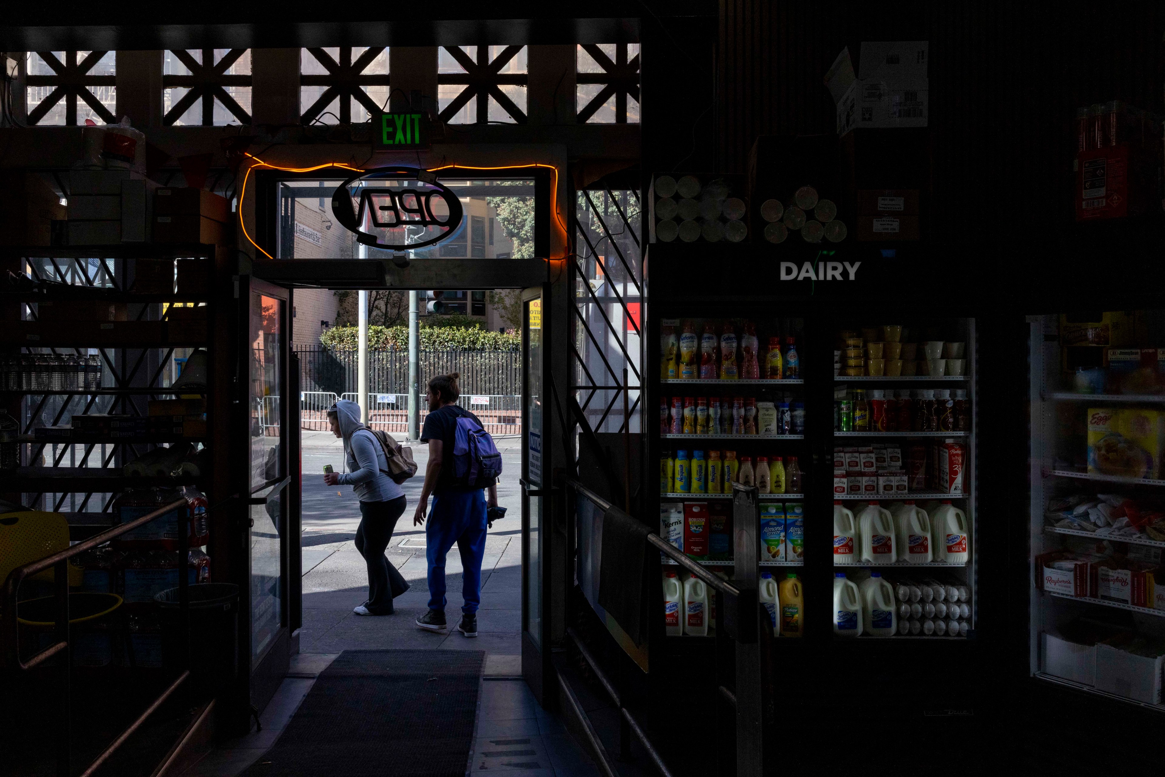 A dimly lit store interior with a glowing &quot;Open&quot; sign above the door. Two people exit, passing dairy shelves filled with milk and drinks.