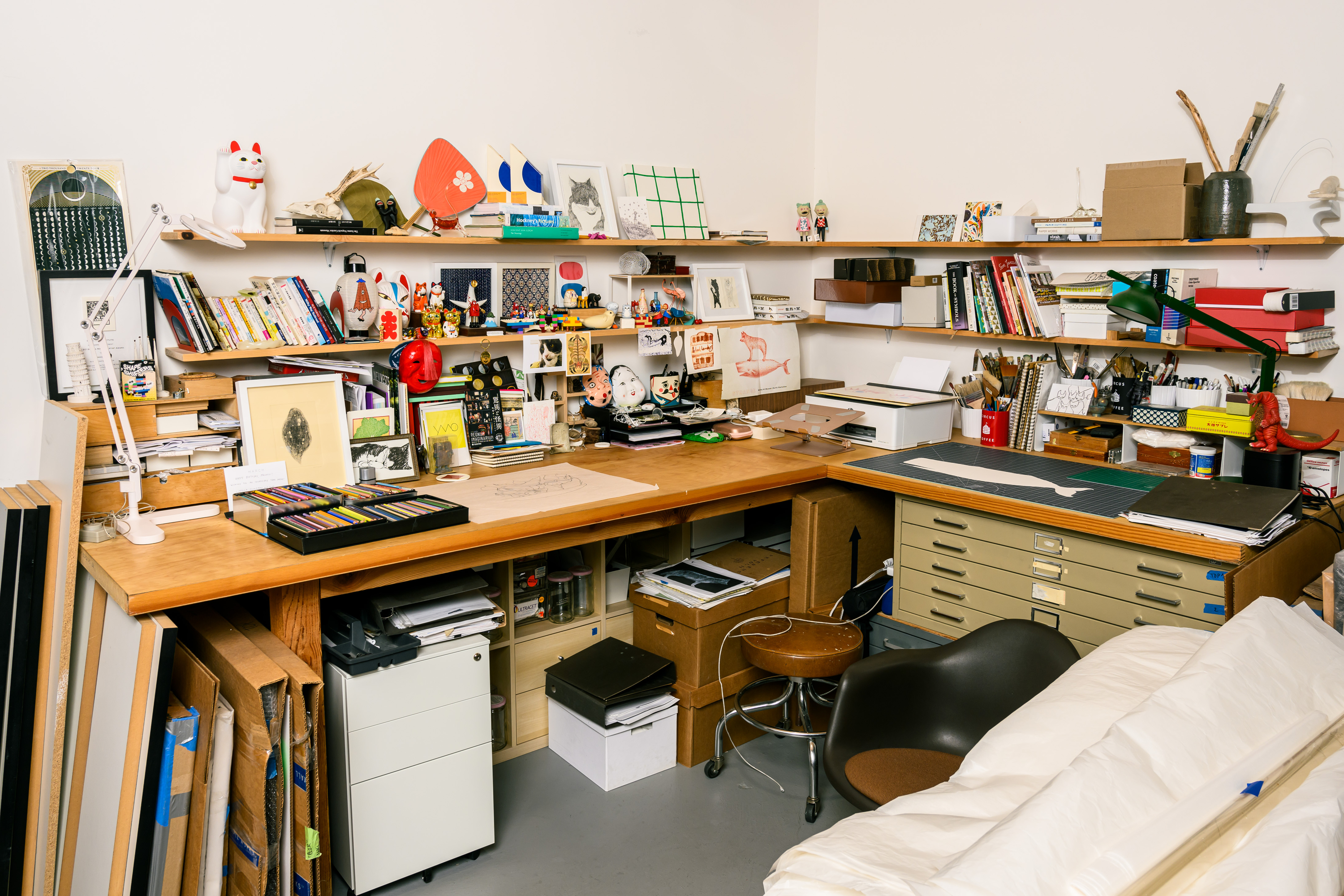 The image shows a cluttered workspace with wooden desks, shelves filled with art supplies, books, colorful art pieces, and various decorative figurines.