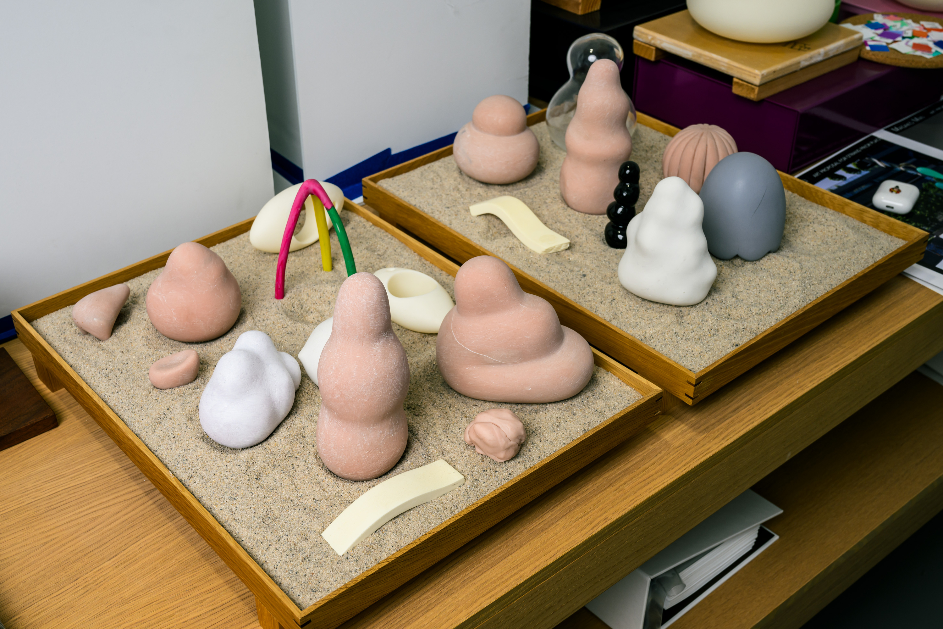 The image shows two wooden trays filled with sand, featuring abstract sculptures in various shapes, sizes, and colors, including pink, white, black, and gray.