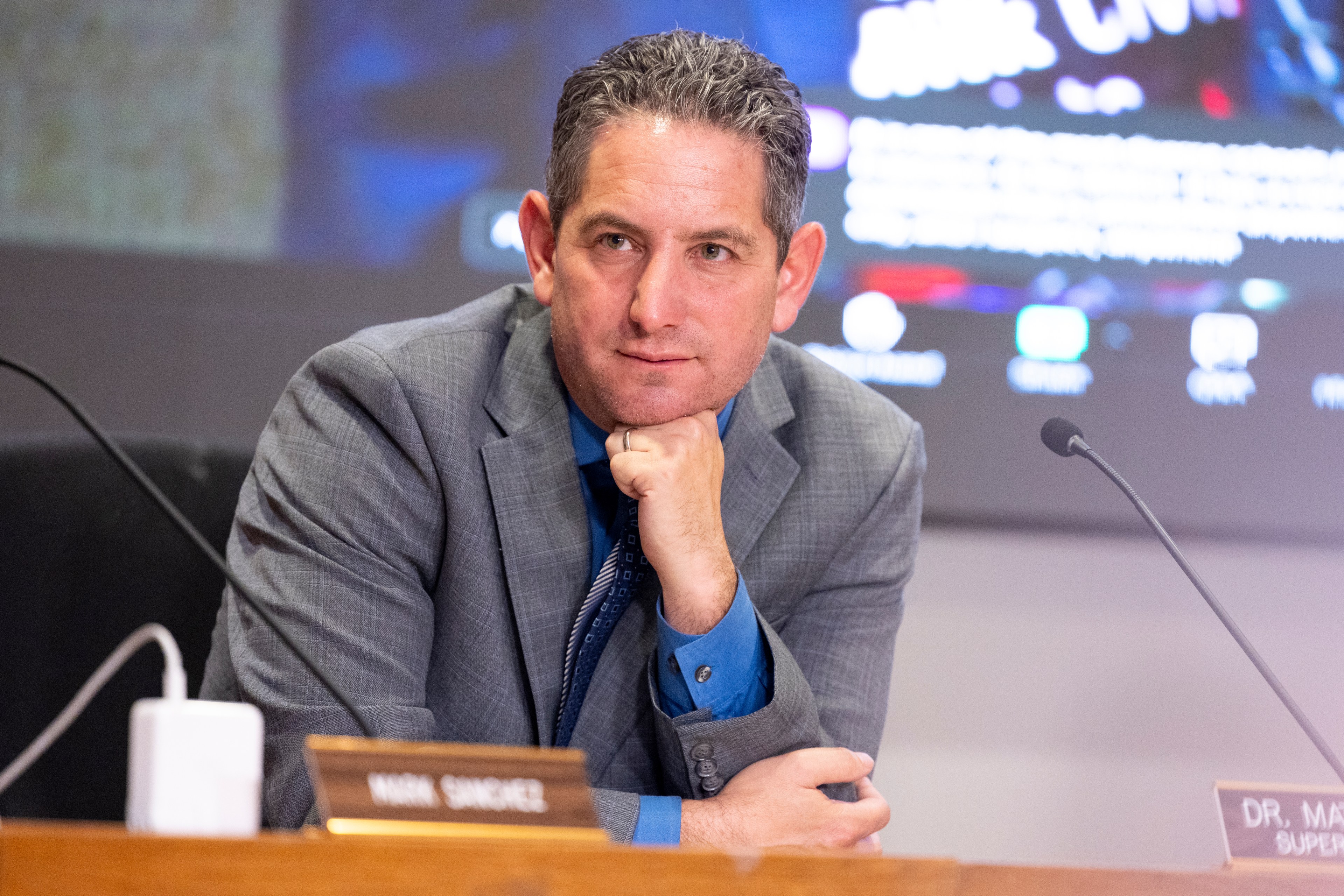 A man in a gray suit and blue shirt sits at a desk with his chin resting on his hand, looking thoughtful. A blurred screen and nameplate are visible behind him.