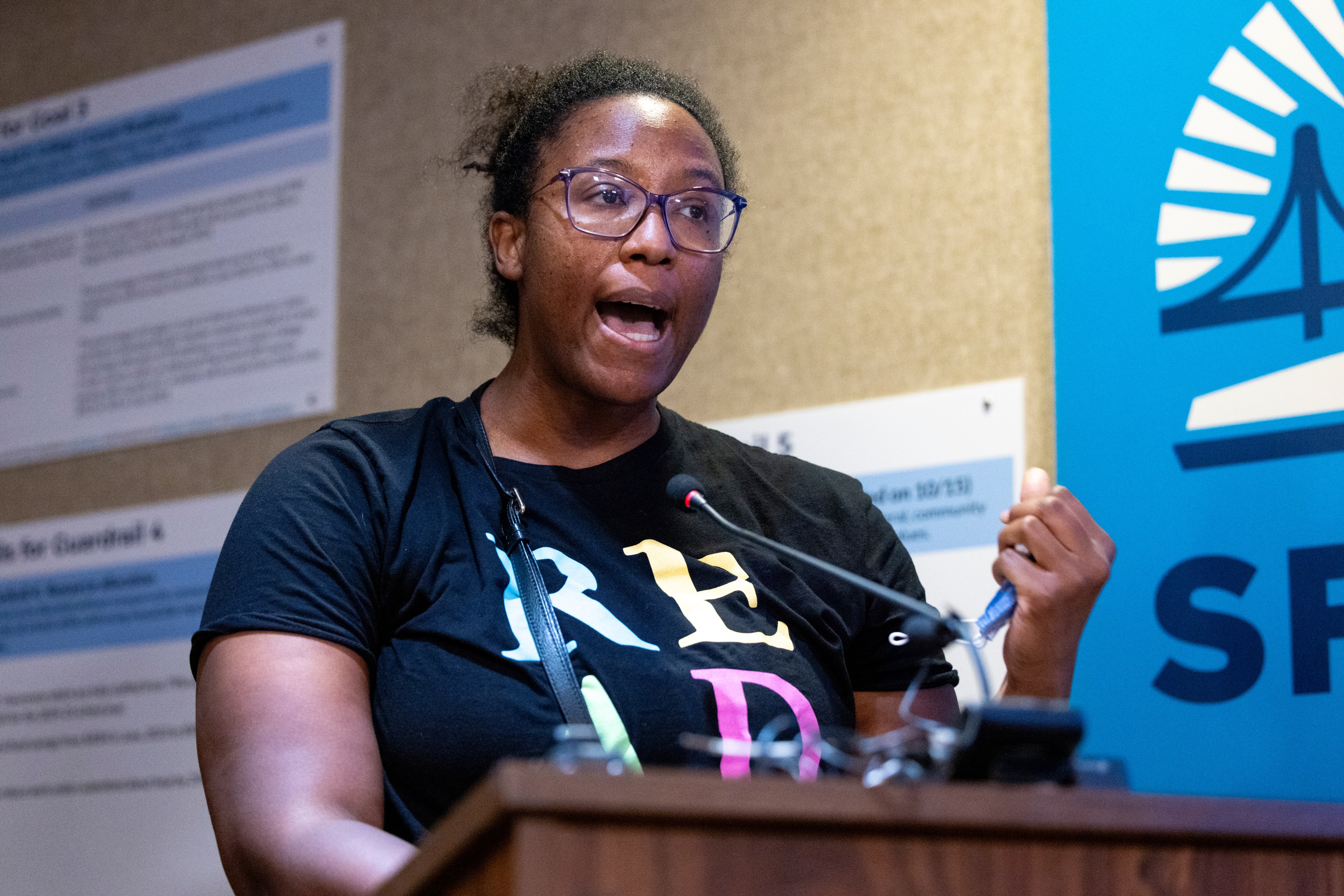 A woman wearing glasses and a black shirt with &quot;READ&quot; in colorful letters speaks passionately at a podium with a microphone. There are informational posters behind her.