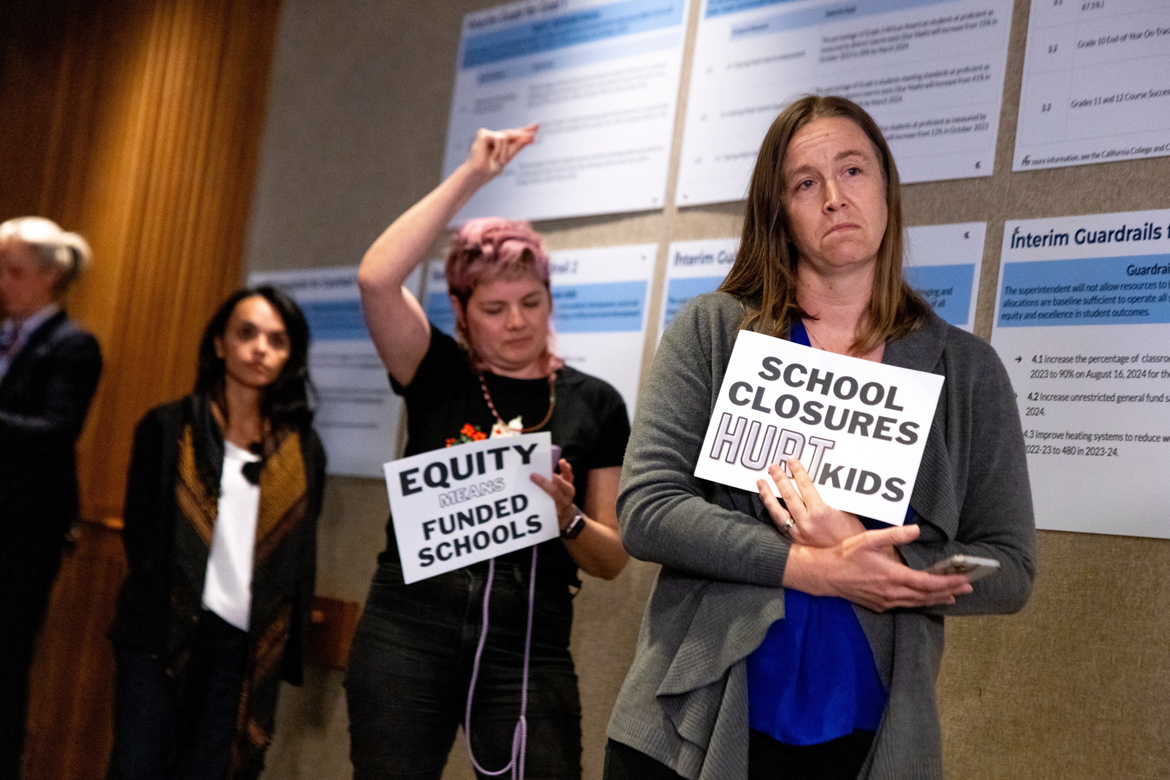 Three people hold signs at a meeting; one reads &quot;School closures hurt kids,&quot; and another reads &quot;Equity means funded schools.&quot; They appear concerned.