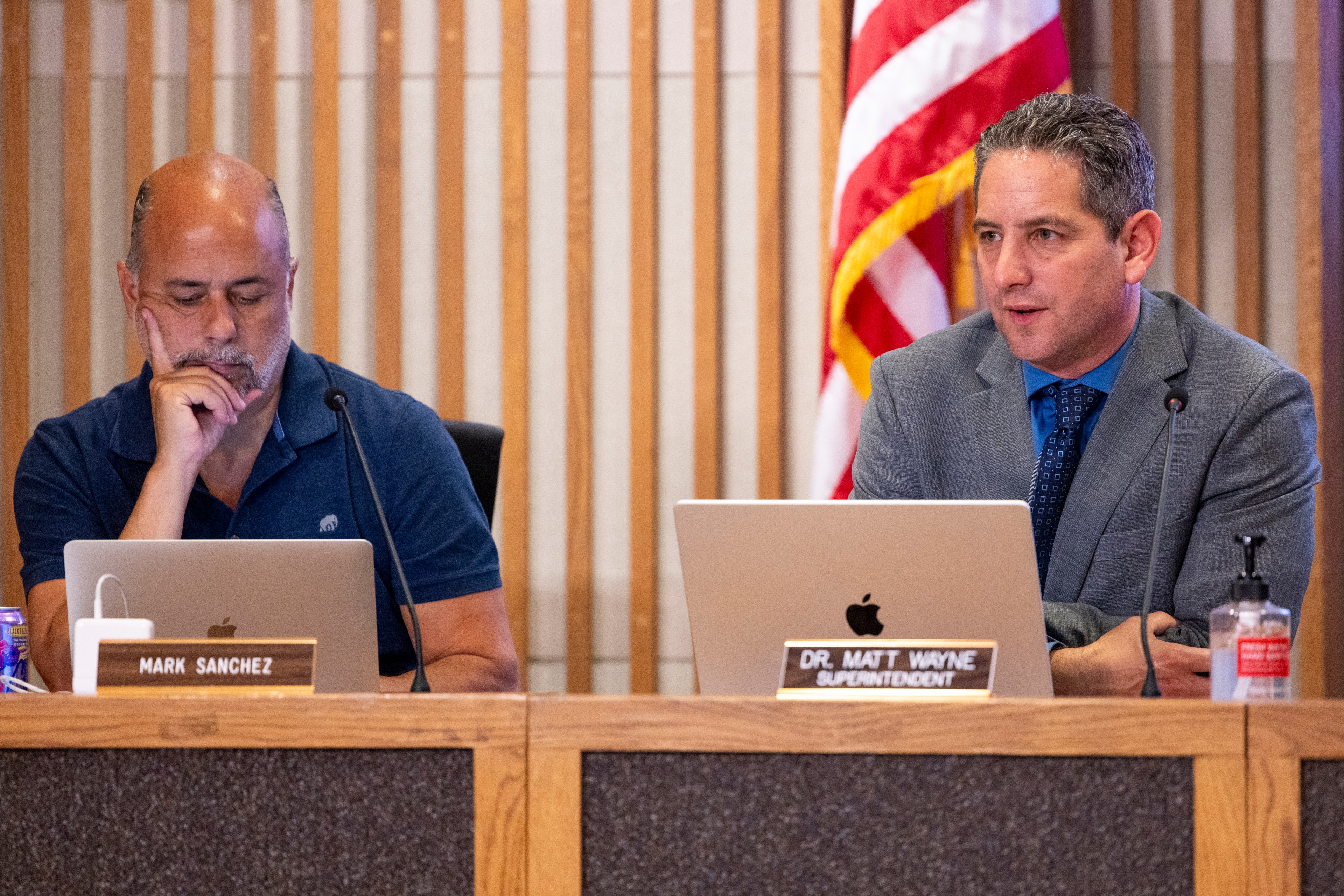 Two men sit at a table with laptops and nameplates. One is focused, and the other is speaking. There's a U.S. flag in the background.