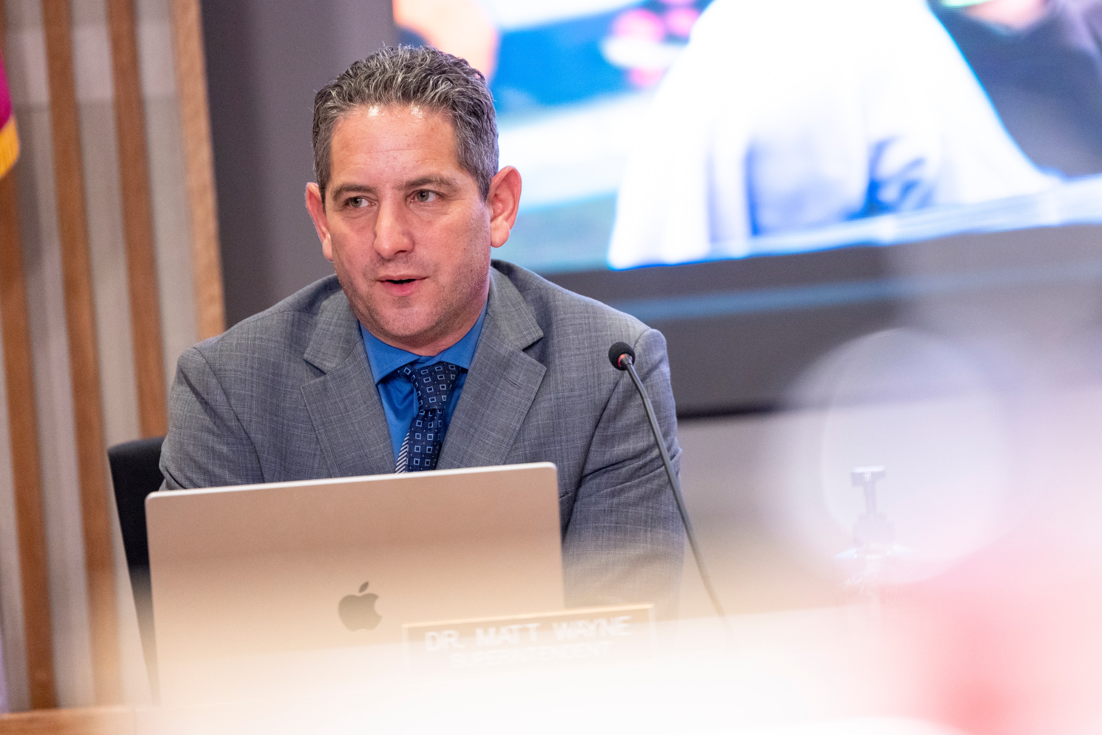 A man in a gray suit and blue shirt is speaking at a microphone. He sits in front of a laptop with a blurred presentation screen behind him.