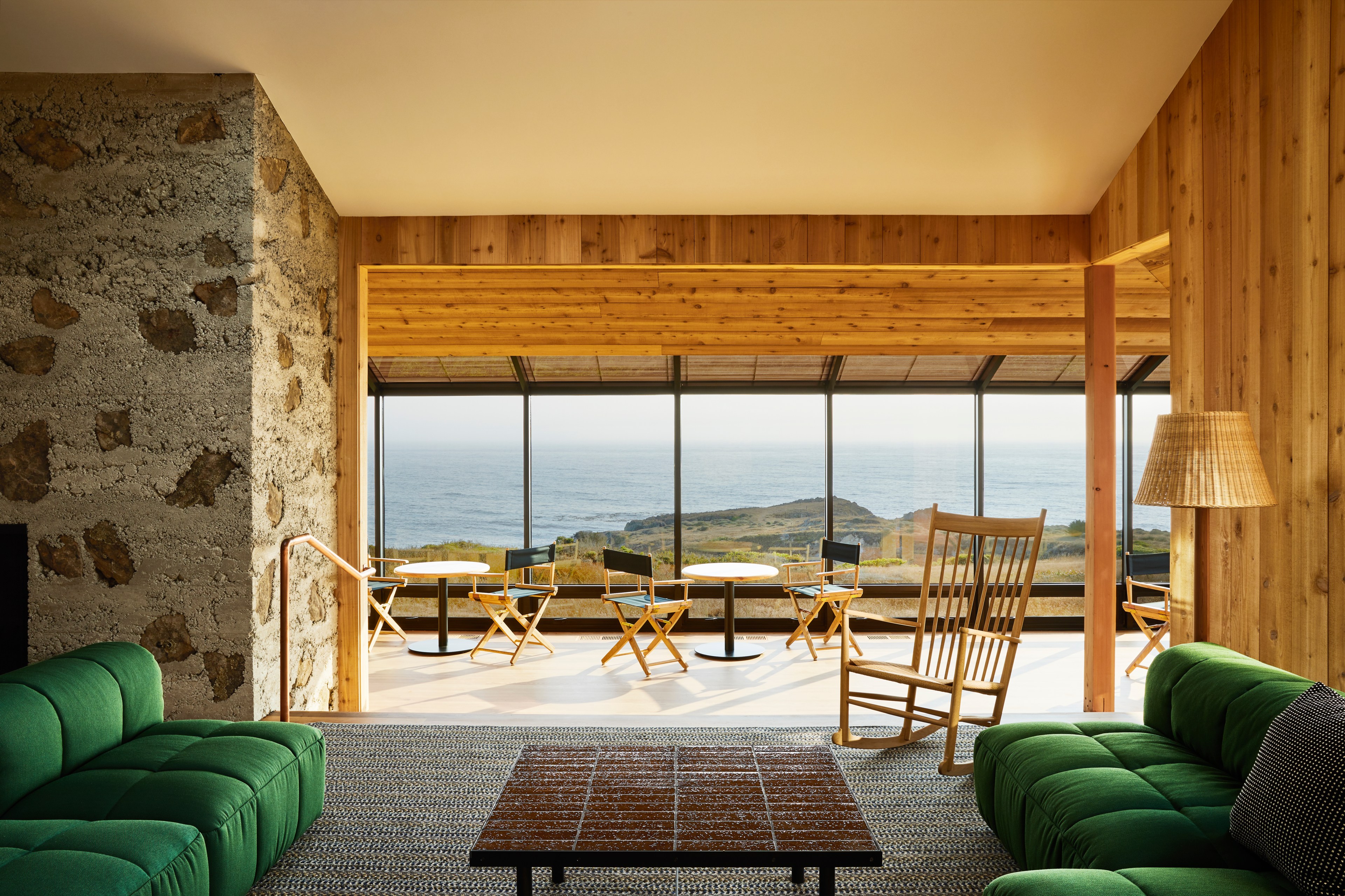 A cozy room with green sofas and a rocking chair overlooks an ocean view through large windows. The walls are wooden, and there’s a stone accent on the left.