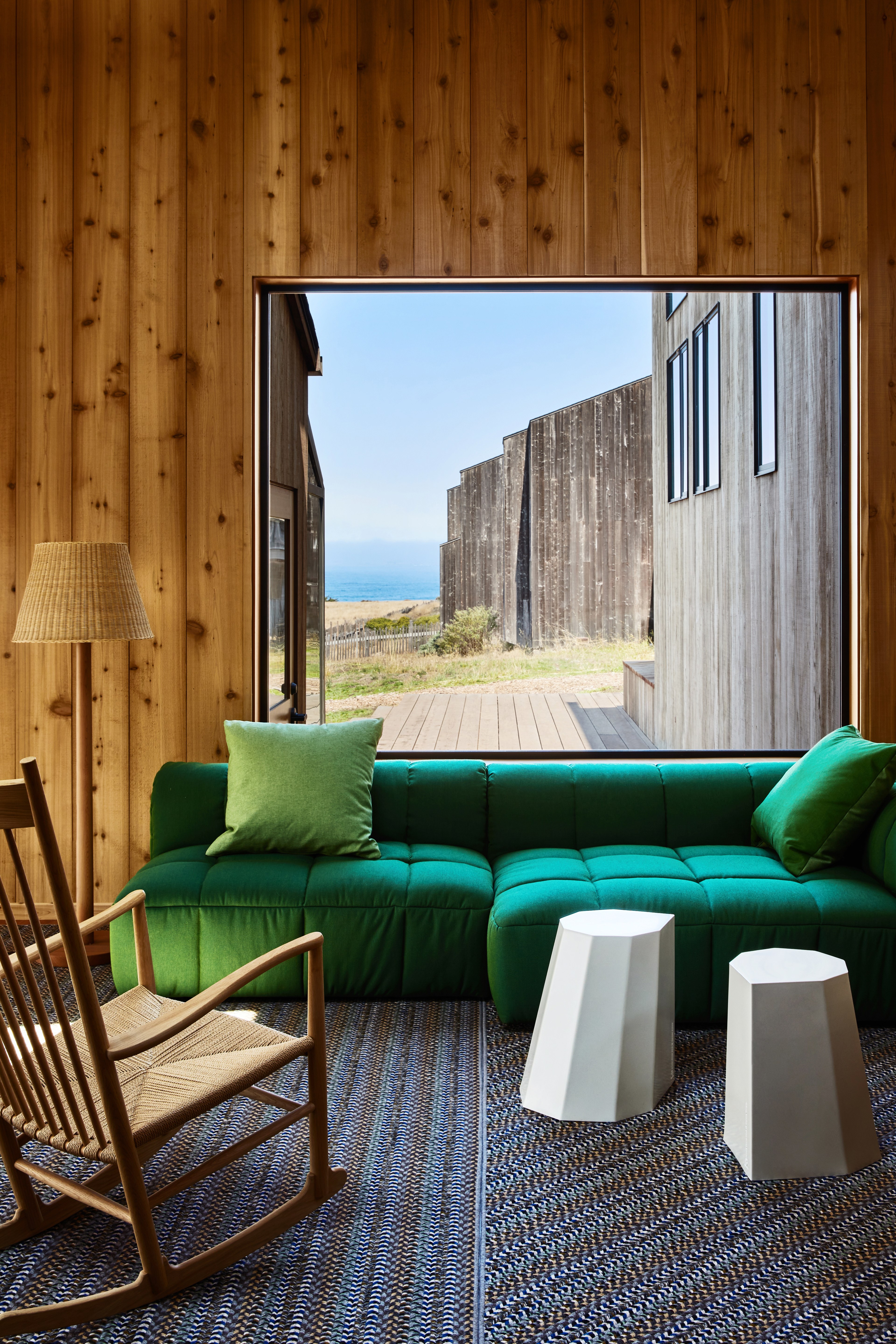 A room features a large window view of the sea, framed by wooden walls. Inside, a green sofa, wicker rocking chair, white stools, and a lamp on a woven rug complete the scene.