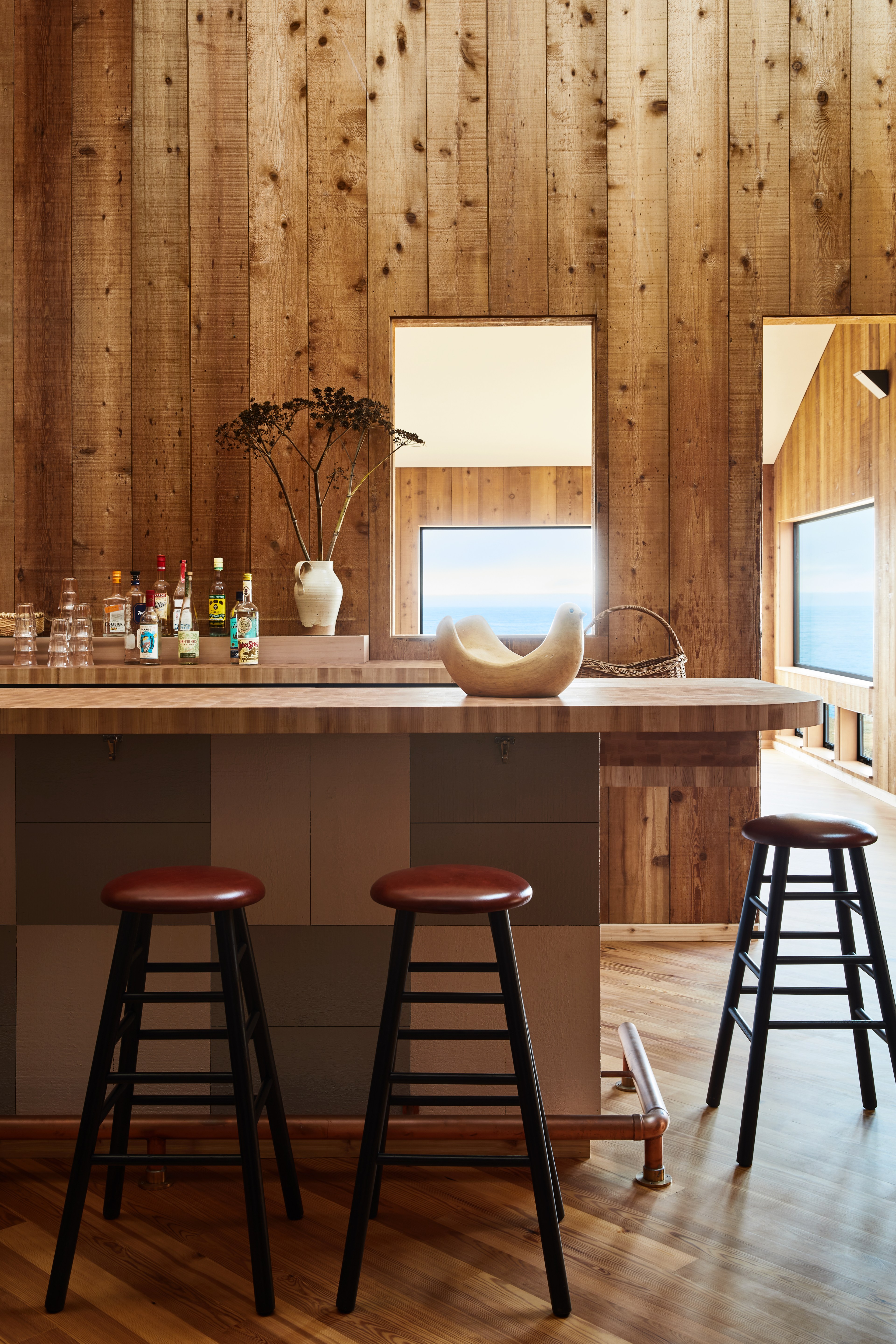 A wooden bar area with tall stools features bottles and a vase on the counter. Large windows reveal a view of the ocean, framed by warm wooden walls.