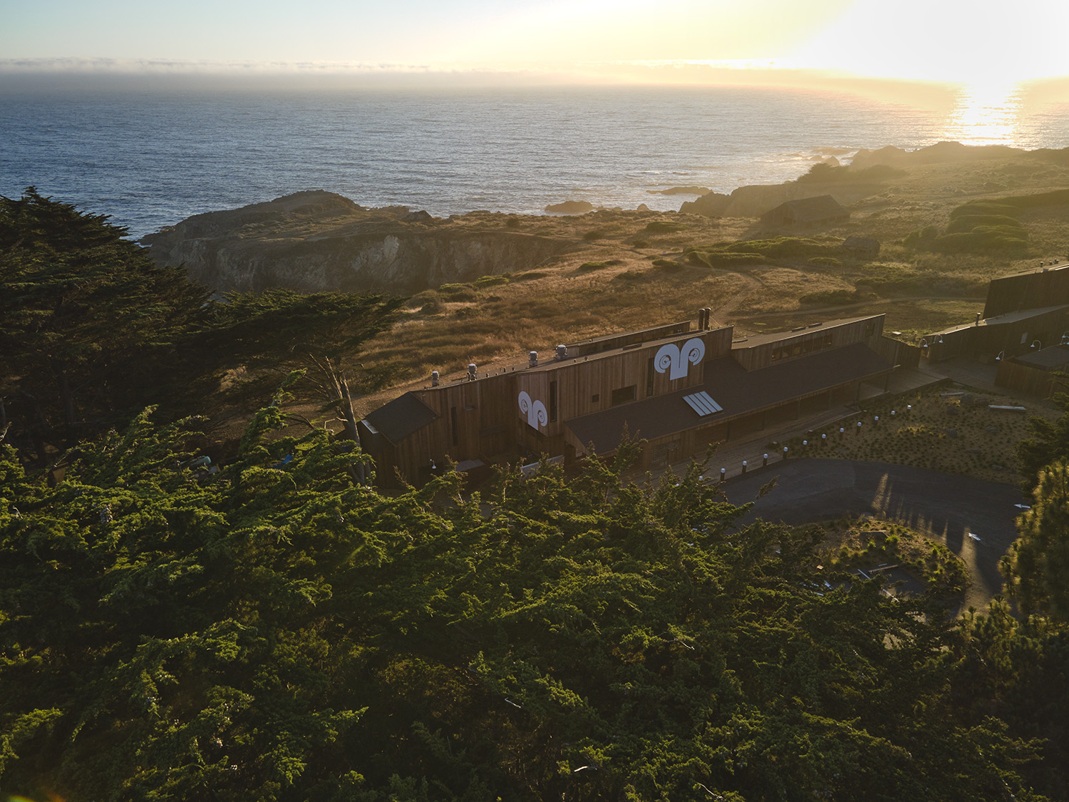 A coastal landscape features a modern wooden building with two circular logos, surrounded by greenery, overlooking cliffs and the ocean at sunset.