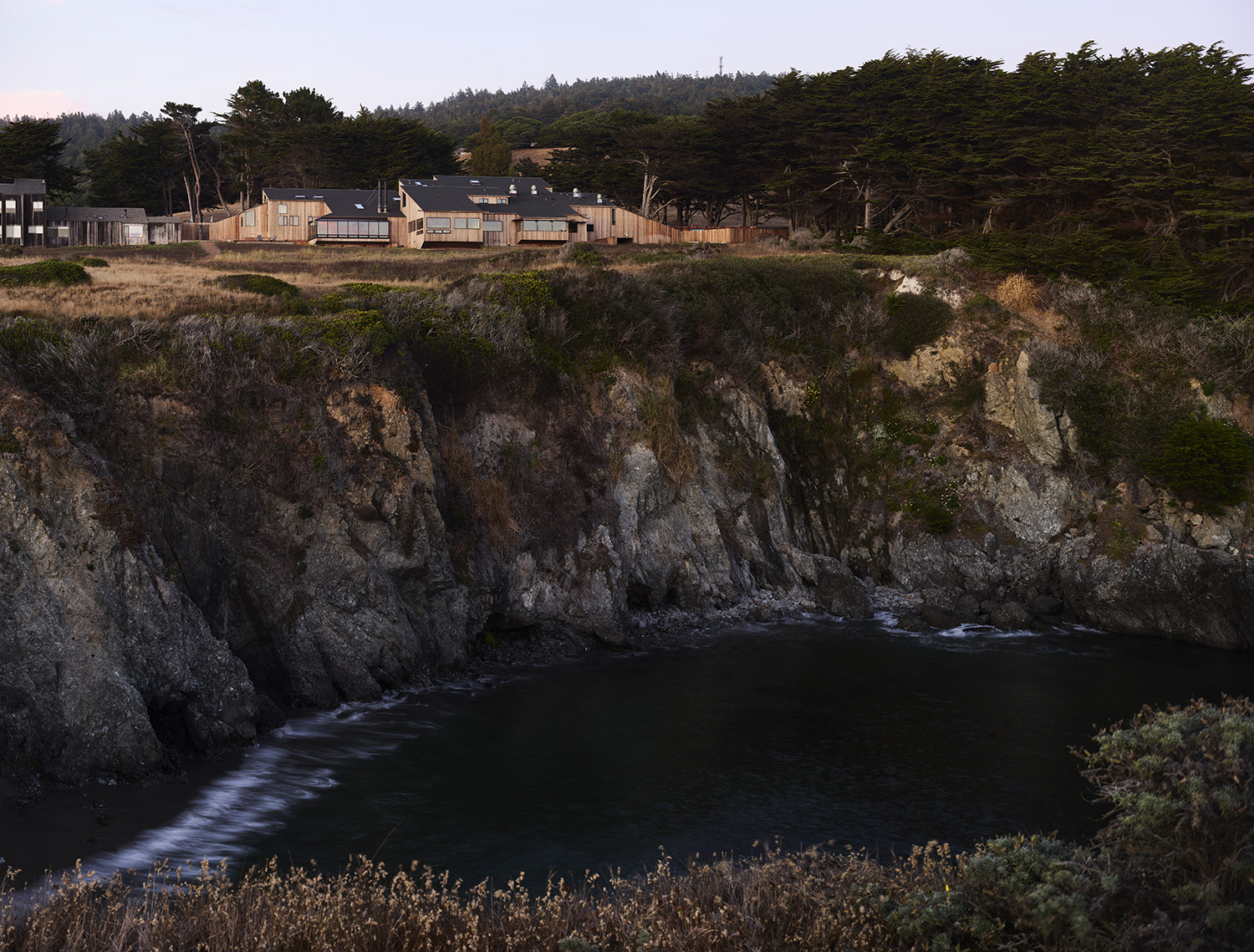 The image shows a rocky coastline with dark waters below. Above the cliffs, there are several wooden buildings surrounded by dense trees and vegetation.