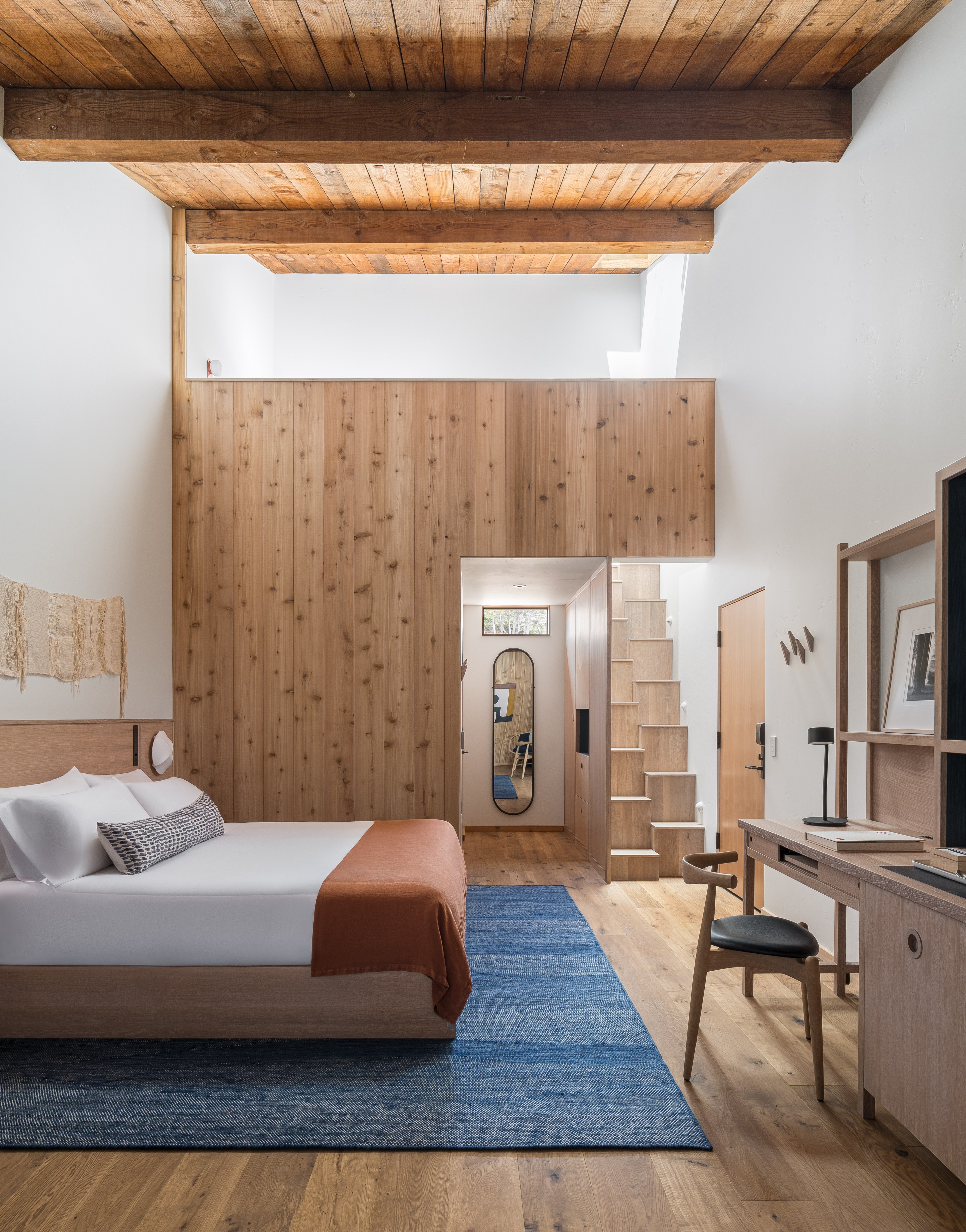 This image depicts a cozy bedroom with a wooden ceiling and walls. A bed with orange and white linens, a blue rug, and a desk with a chair are visible.