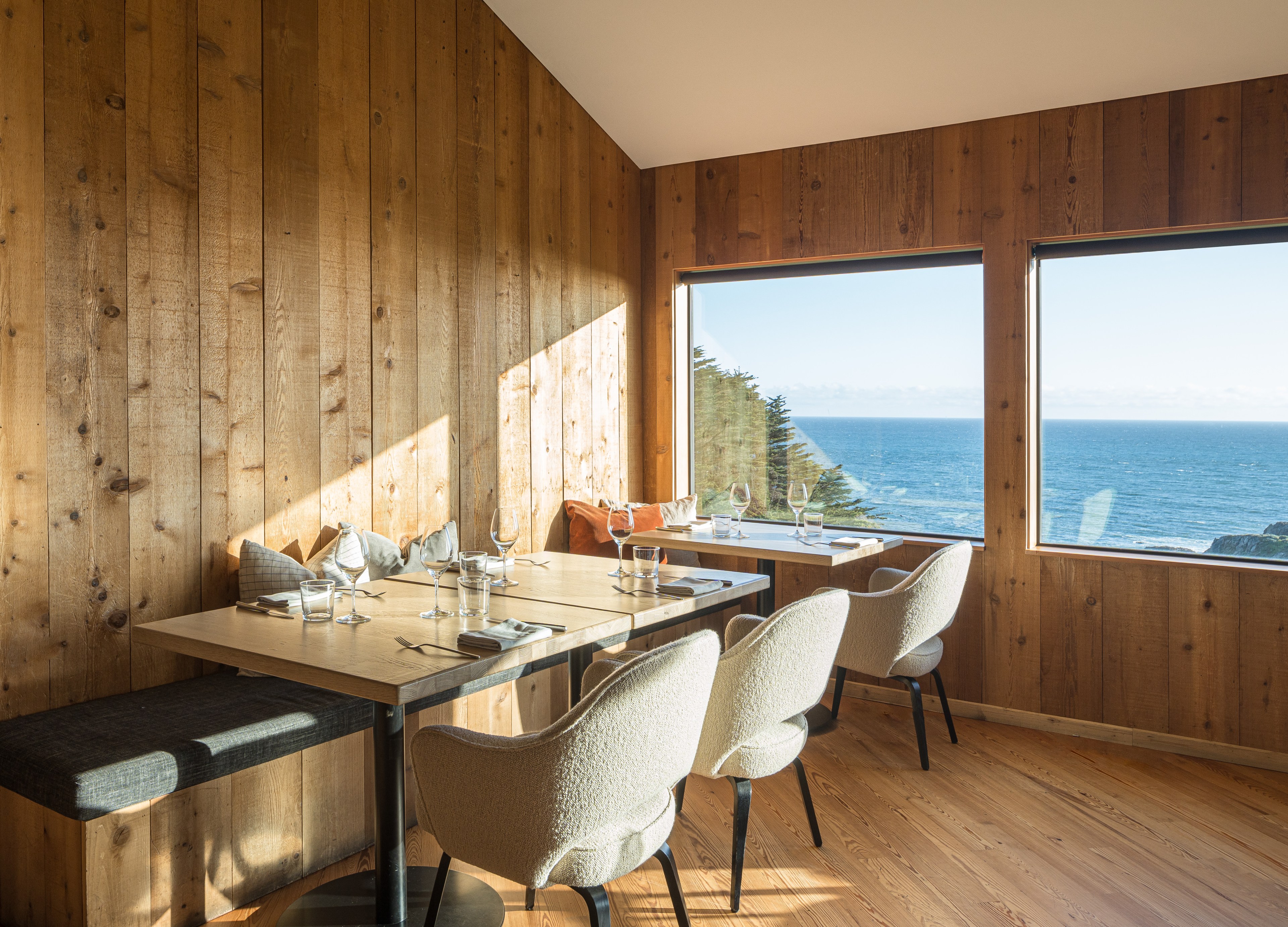 A cozy dining area with wood walls and two tables set with glasses and cutlery. Large windows offer a view of the ocean and trees.