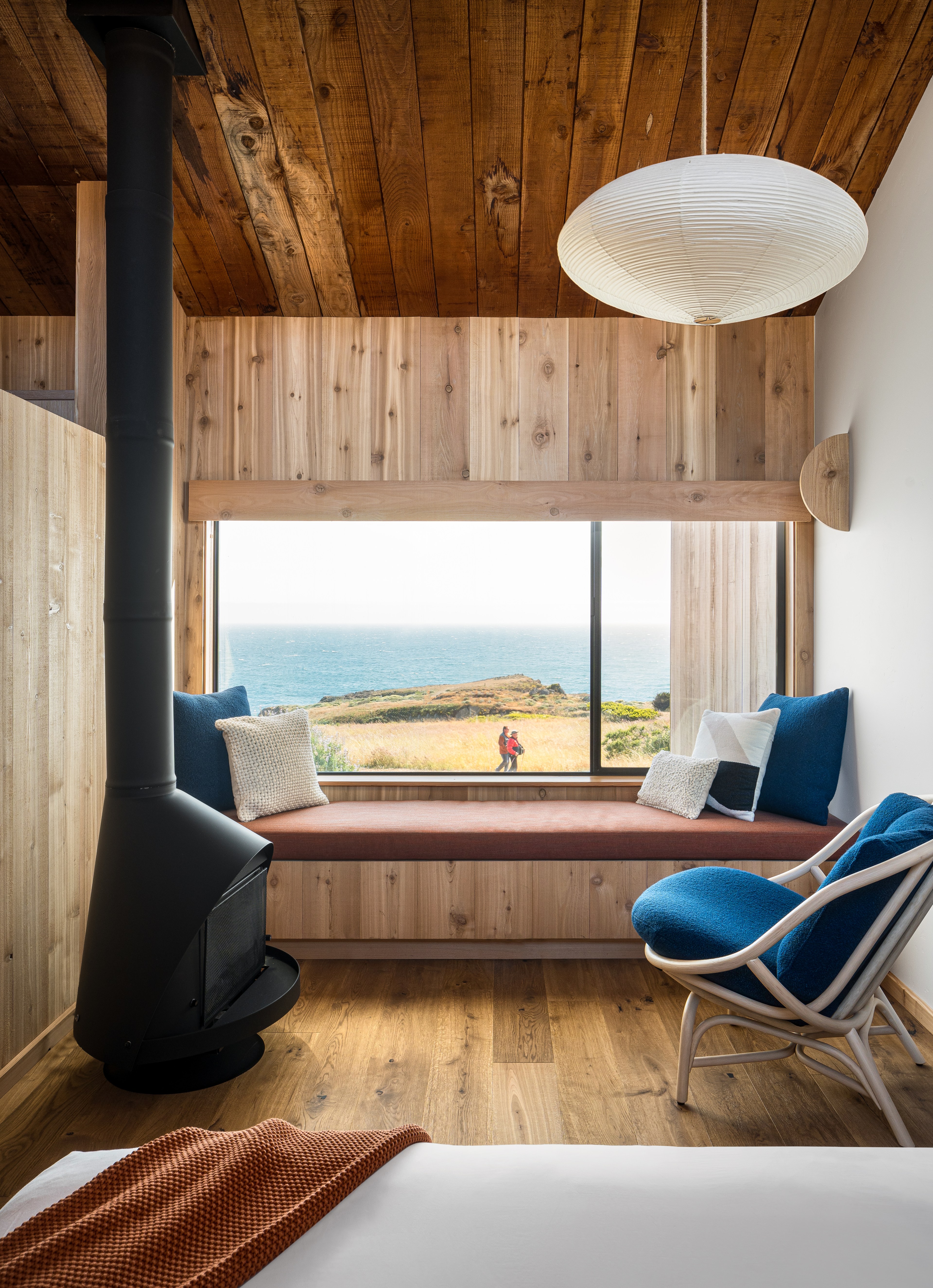 A cozy room with wooden walls and ceiling features a large window with ocean views, a bench seat with cushions, a hanging lamp, and a modern black fireplace.