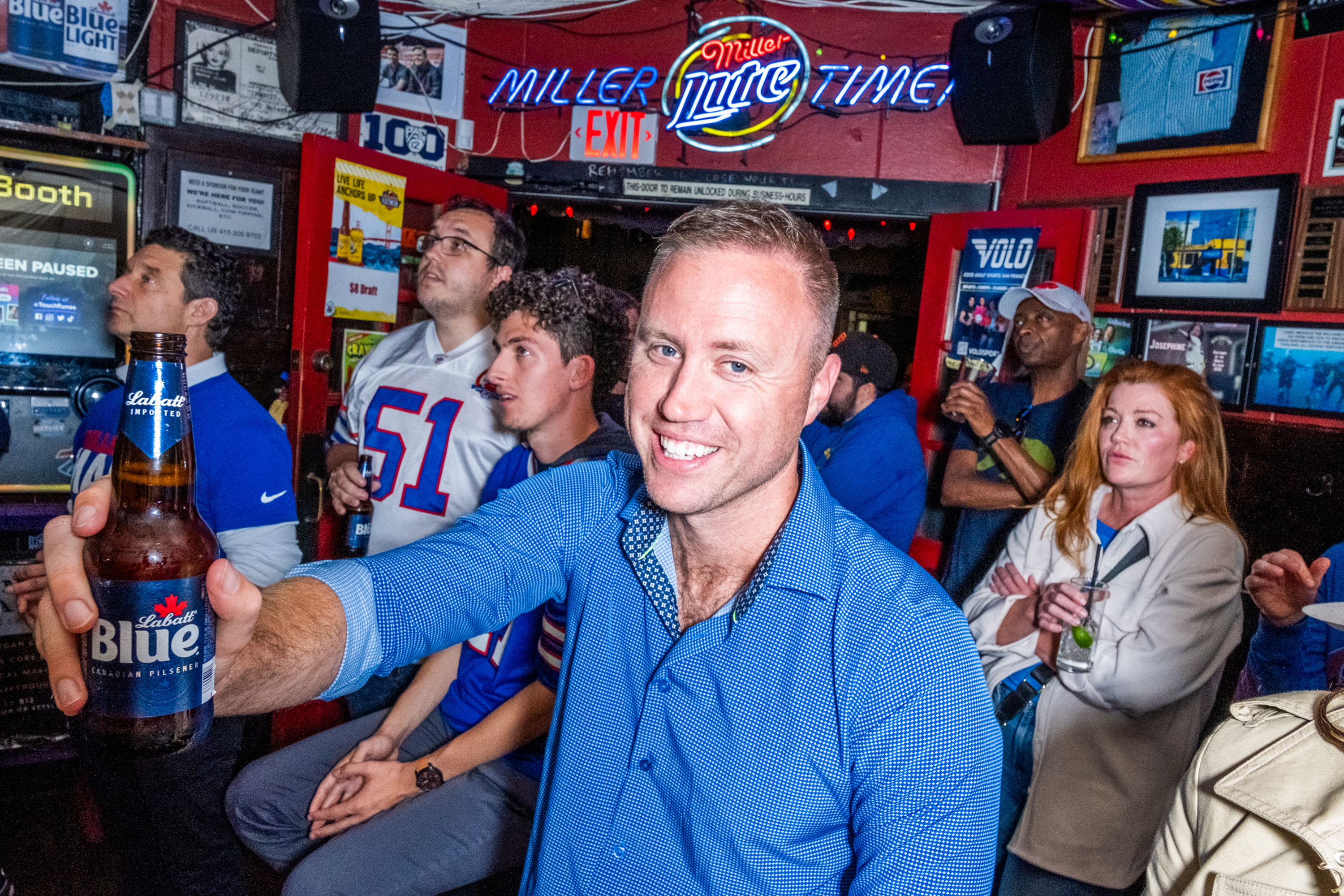 A man in a blue shirt holds a Labatt Blue beer smiling in a busy bar. People in sports jerseys watch a TV intently, surrounded by sports memorabilia and neon signs.